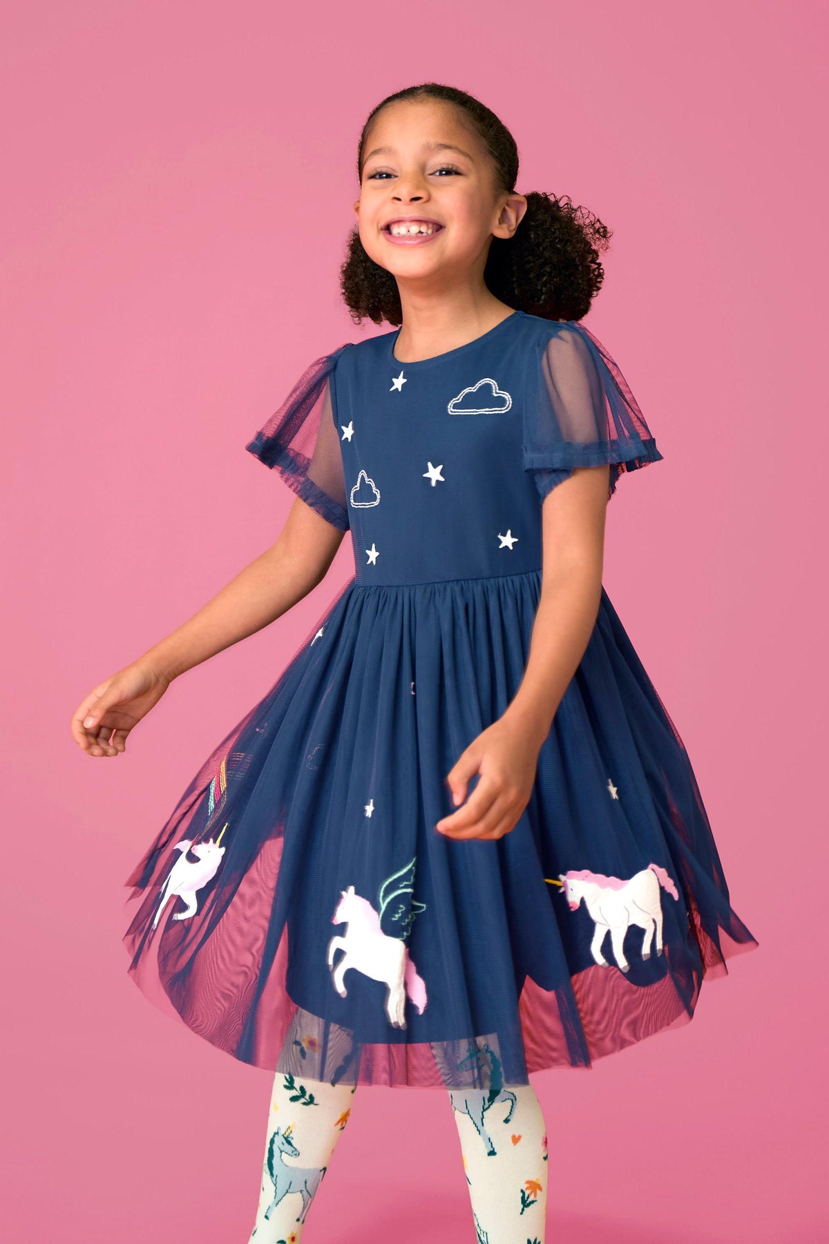 A smiling girl wearing a navy blue Frugi party dress with unicorn and star patterns against a pink background.