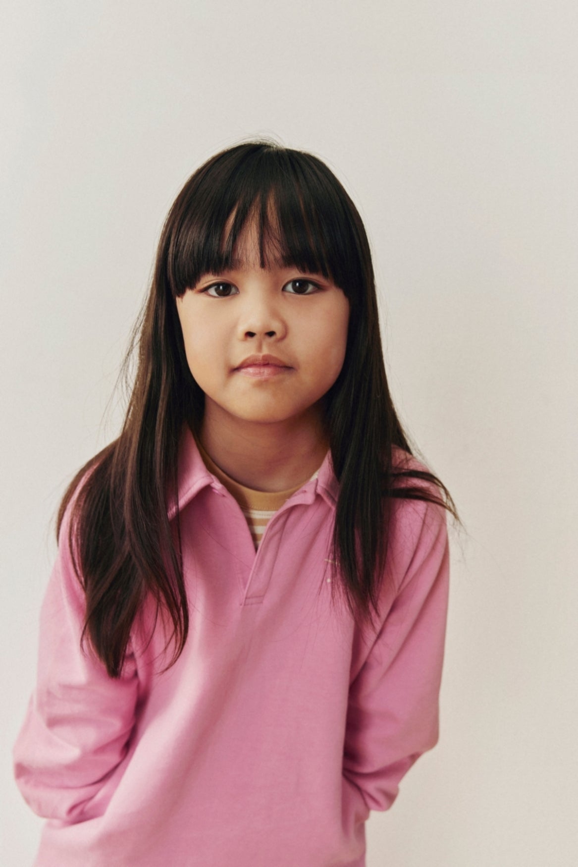A young girl with long dark hair wearing a pink long-sleeve polo shirt, photographed against a plain light background.