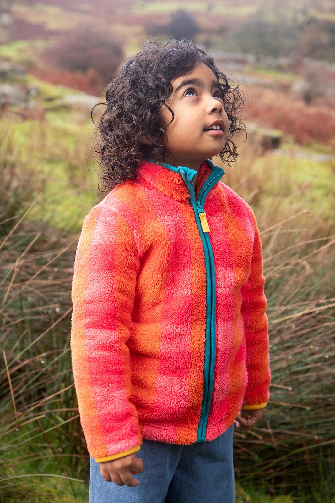 A young girl wearing a colourful Frugi recycled polyester fleece jacket outdoors in a natural landscape.
