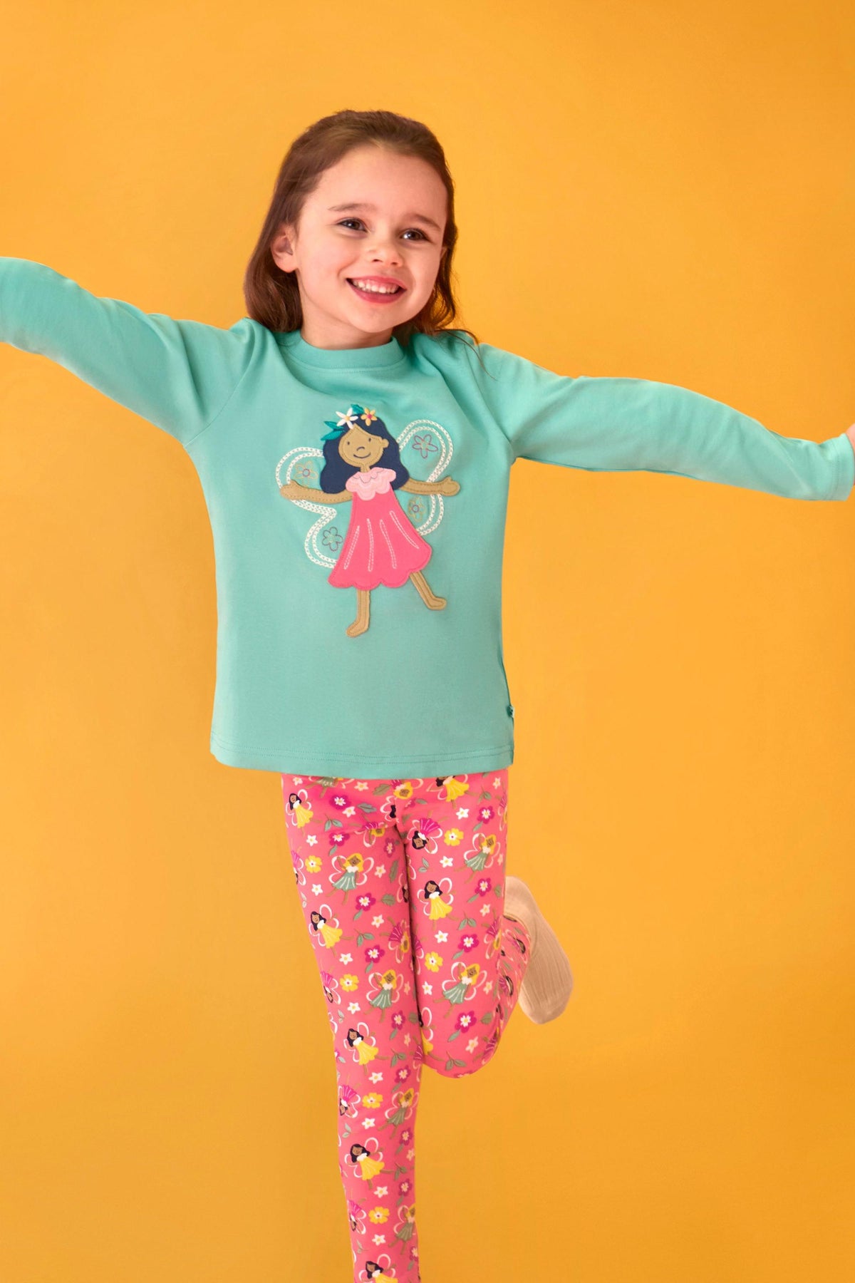 A happy young girl wearing a teal Frugi fairy print organic cotton top jumping against a yellow background.