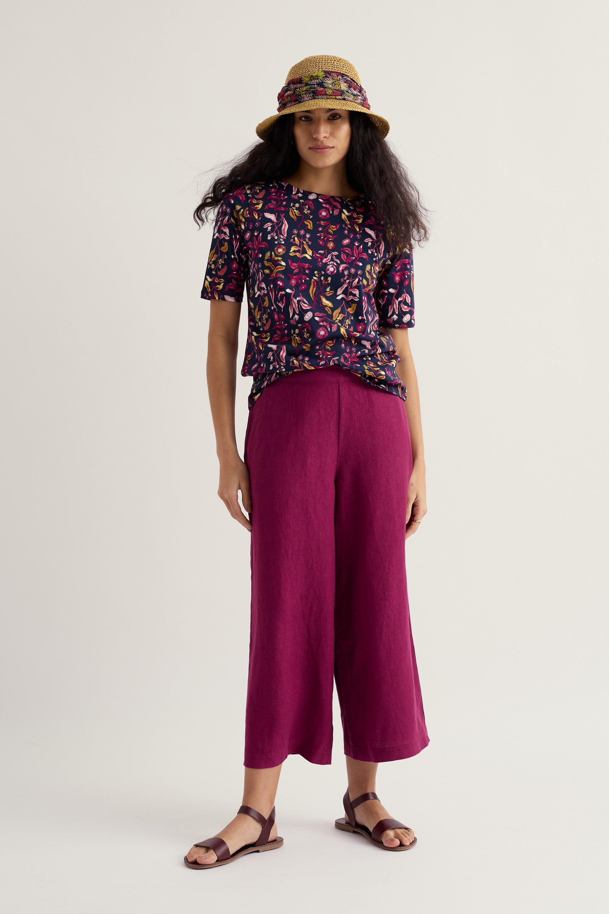 Woman wearing a patterned Seasalt Cornwall top and magenta pants with a hat on a white background
