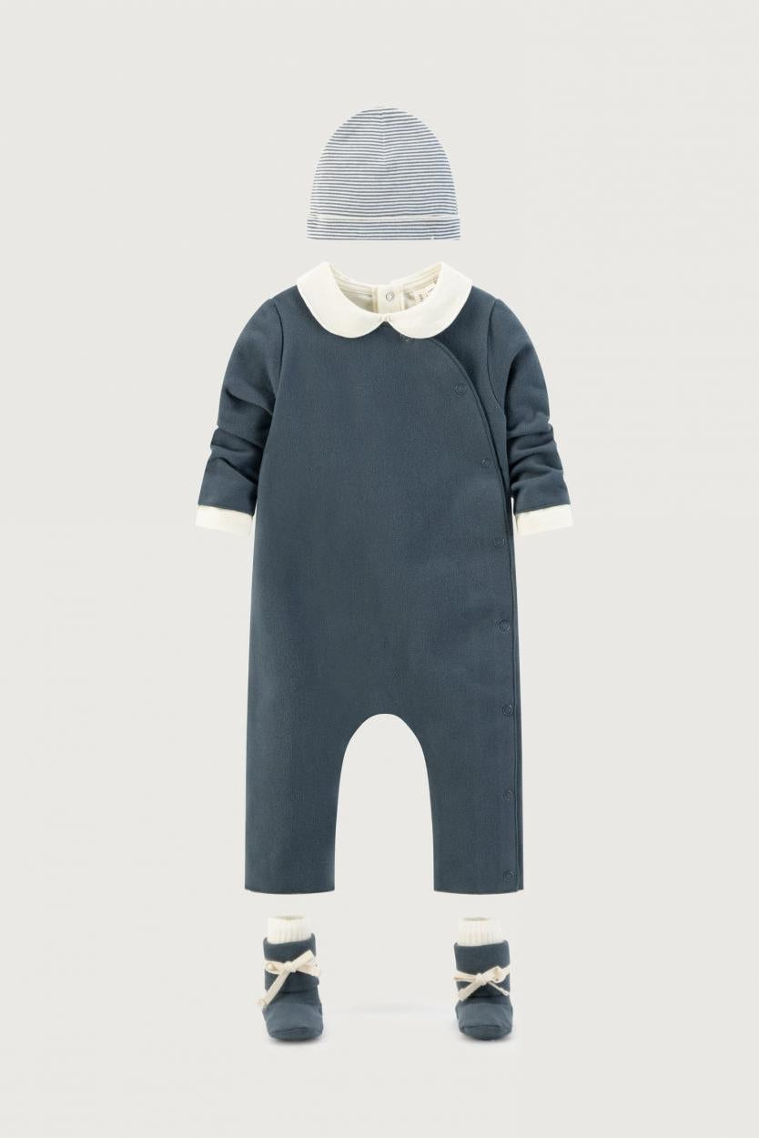 A baby outfit set featuring a dark slate blue one-piece jumpsuit with a cream collar, a matching striped bonnet, cream socks, and blue booties, displayed on a neutral light gray background.