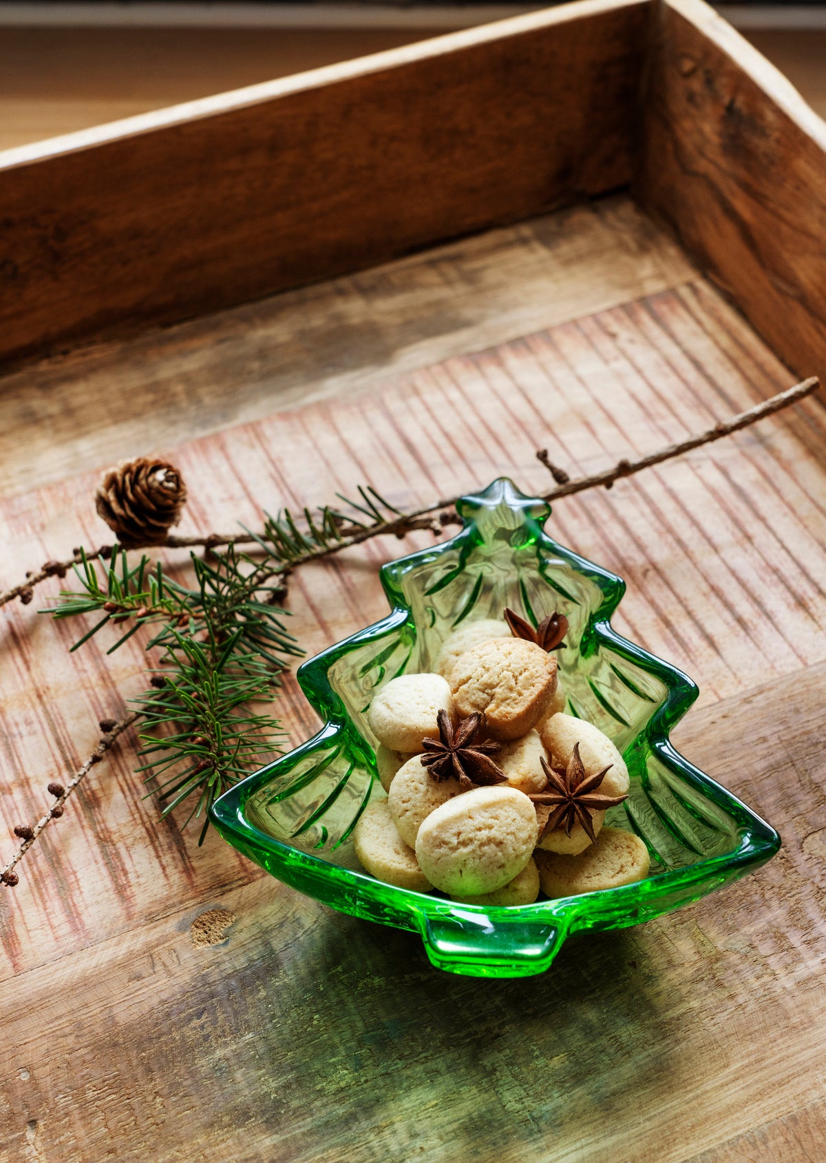 A green glass christmas tree dish filled with cookies and star anise on a rustic wooden tray.
