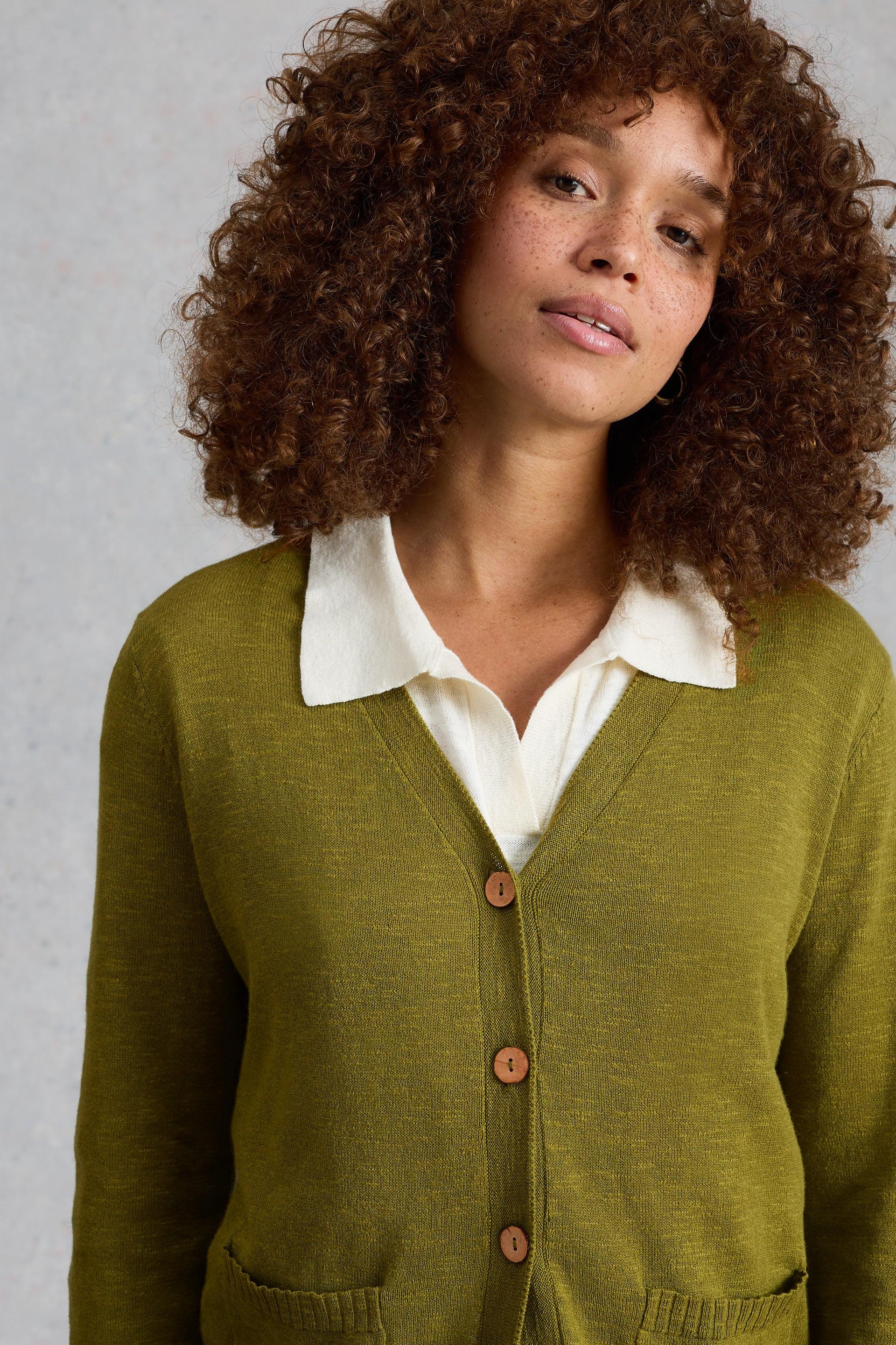 Woman wearing a green White Stuff cardigan over a white shirt against a gray background