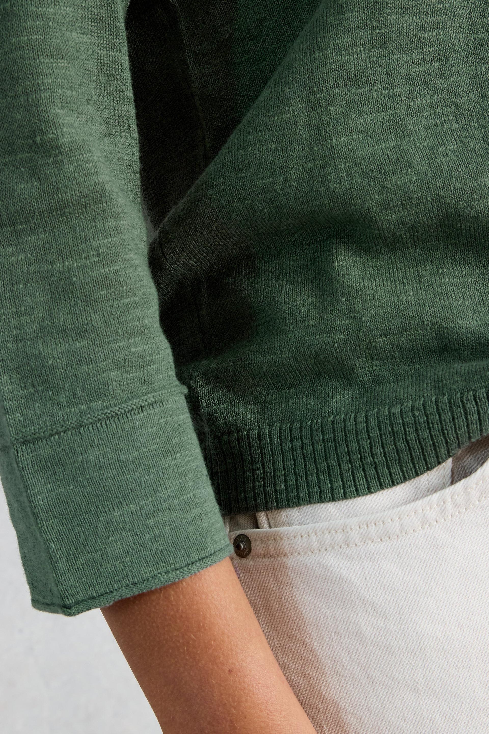 Close up of a person wearing a green organic cotton v-neck sweater paired with white jeans.