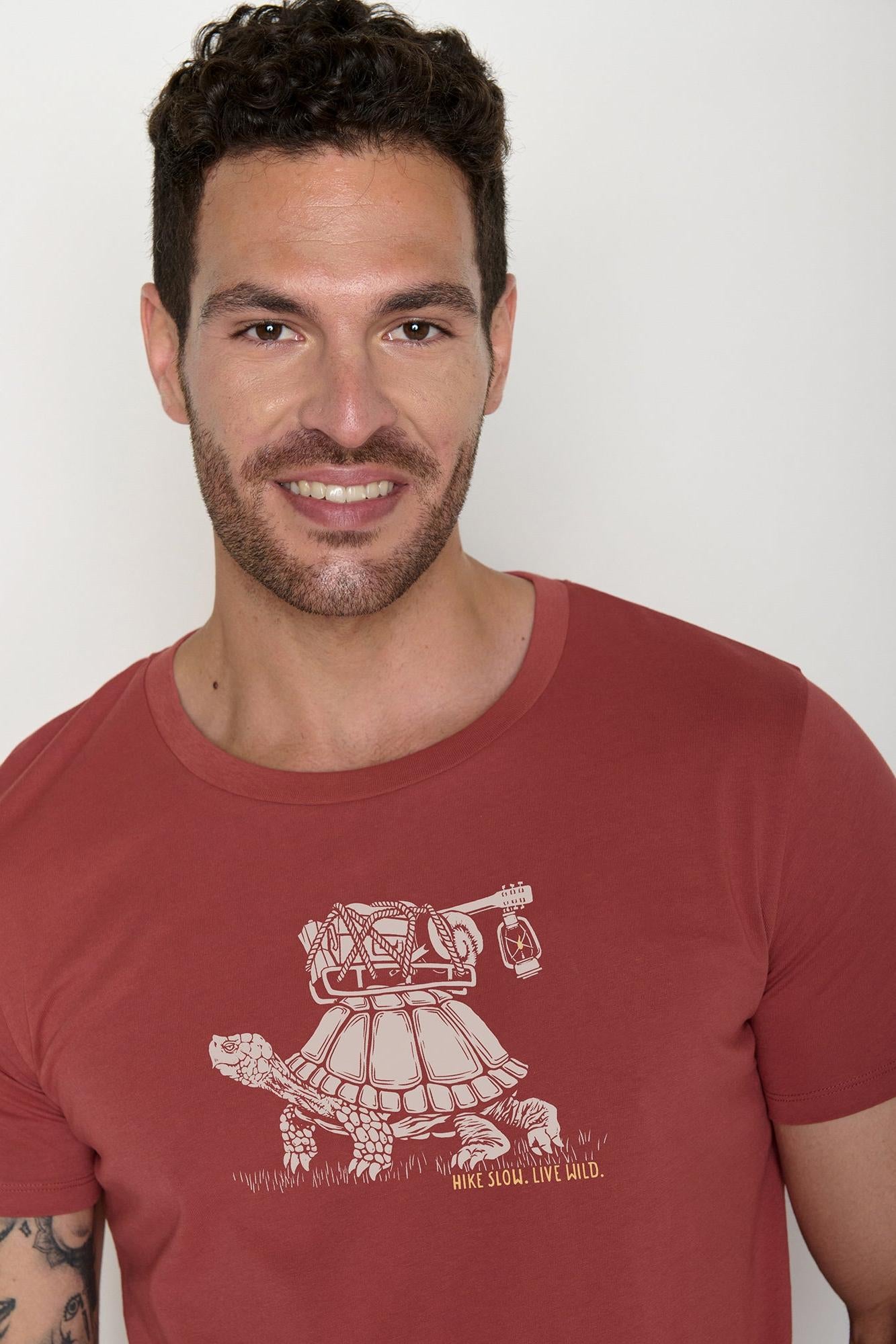 Man wearing a red Greenbomb t-shirt with a graphic turtle design on a plain background