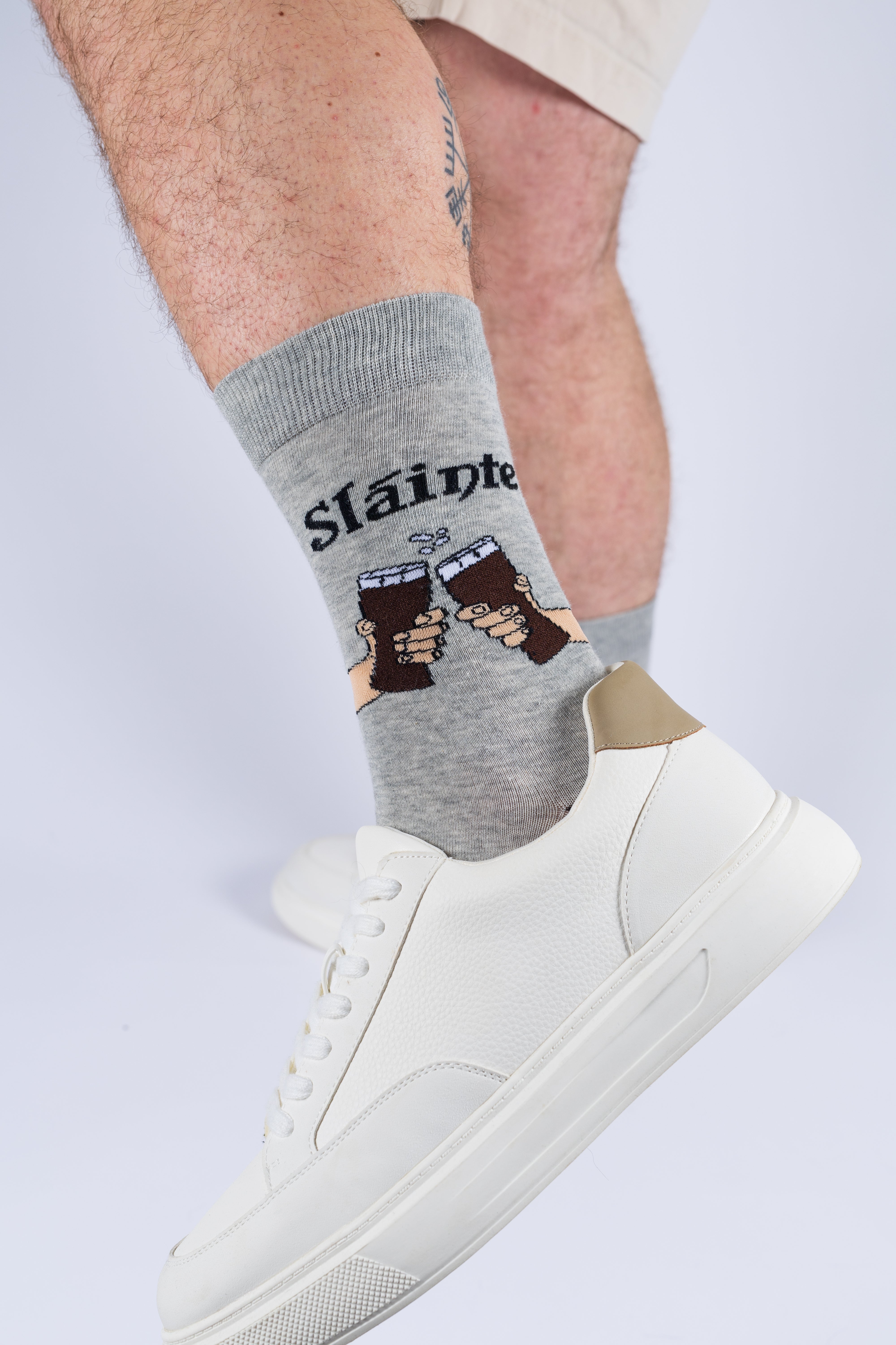 A person wearing grey bamboo socks with a Sláinte toast print and white sneakers against a white background