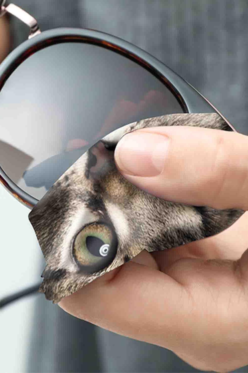 A hand using a microfiber cloth featuring a cat print to clean the dark lens of a pair of sunglasses.