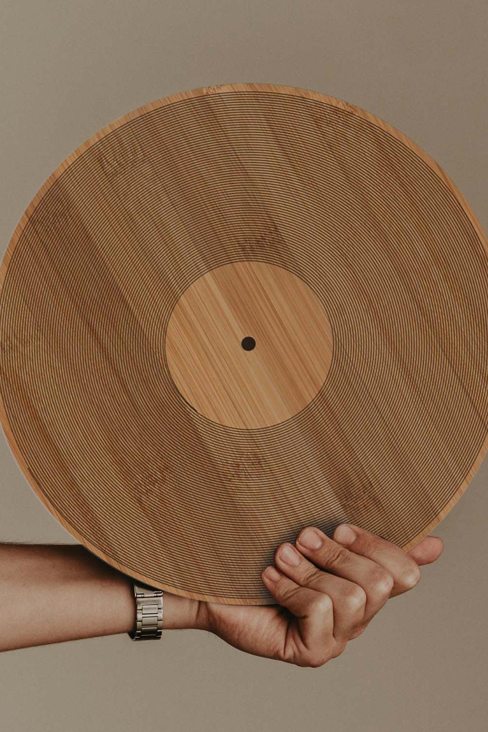 A hand holds a large round wooden board with a natural bamboo grain pattern and a central circular cutout, set against a solid taupe background.
