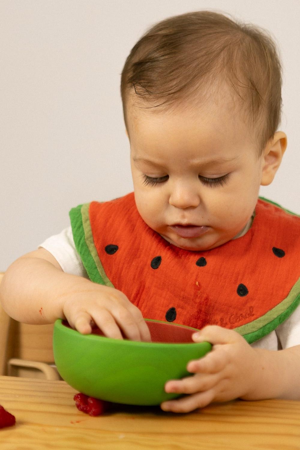 Baby eating and wearing a watermelon-themed bib playing with a green bowl on a wooden table.