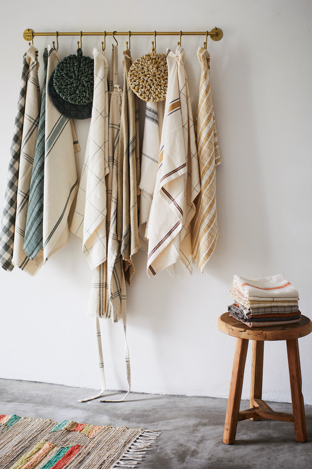 A collection of various patterned cotton towels hanging on a brass rail with a stack on a wooden stool.