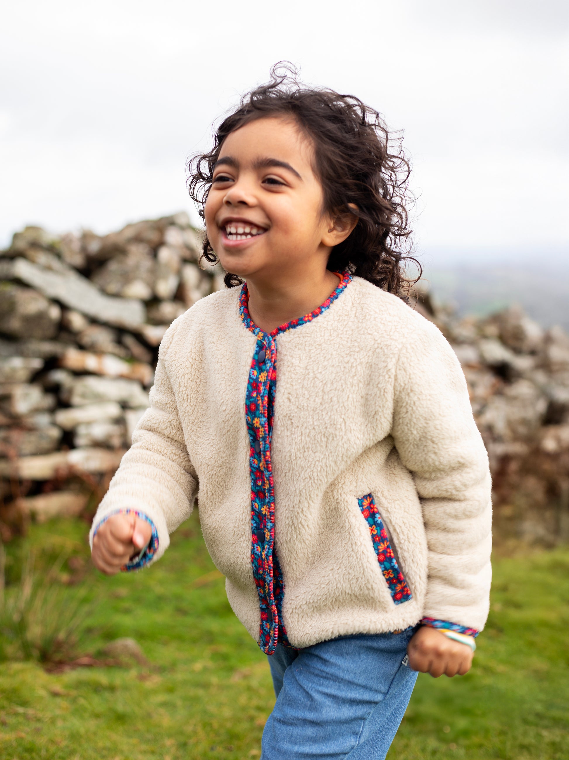 A joyful young girl running outdoors wearing a cream-colored Frugi recycled polyester fleece jacket.