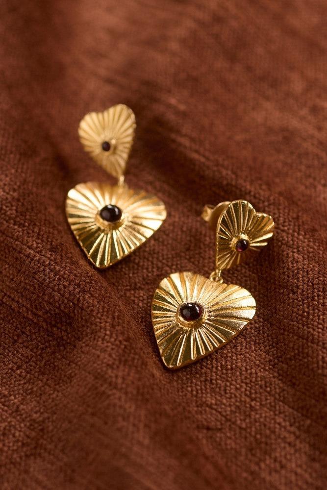 Pair of heart-shaped gold-plated garnet gemstone earrings on a textured brown fabric background.