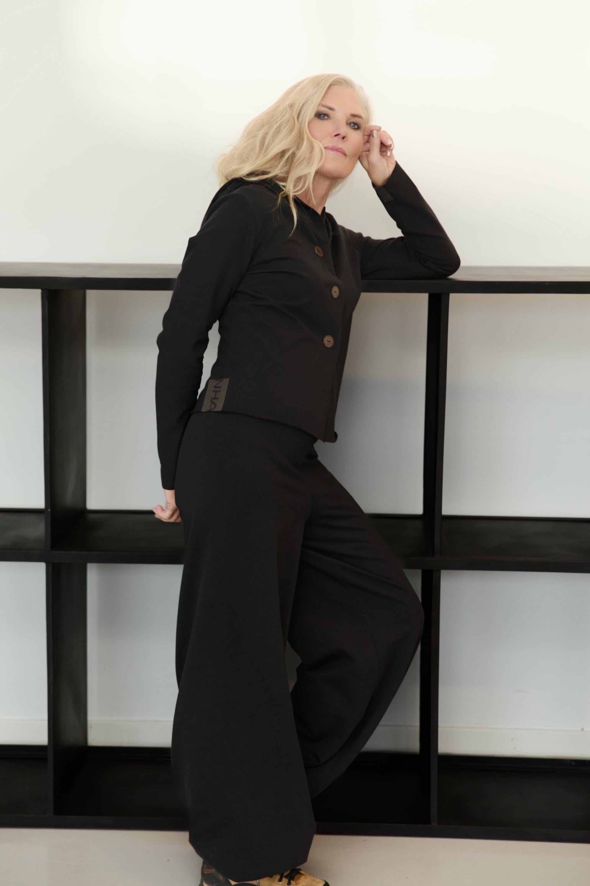 Blonde woman wearing a Henriette Steffensen black organic cotton hoodie standing against a black shelf.