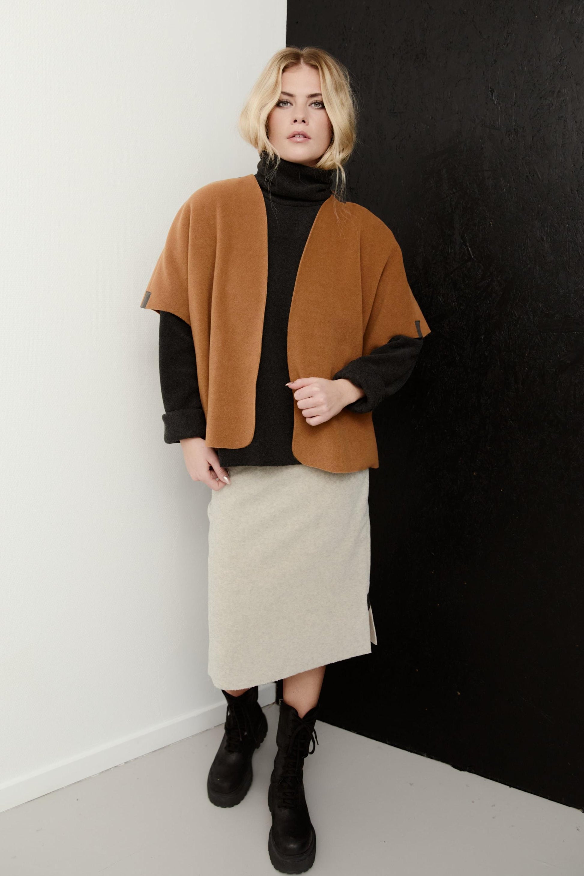 A woman models a cape jacket in camel brown, worn over a black turtleneck and a light beige skirt, standing against a split white and black wall.