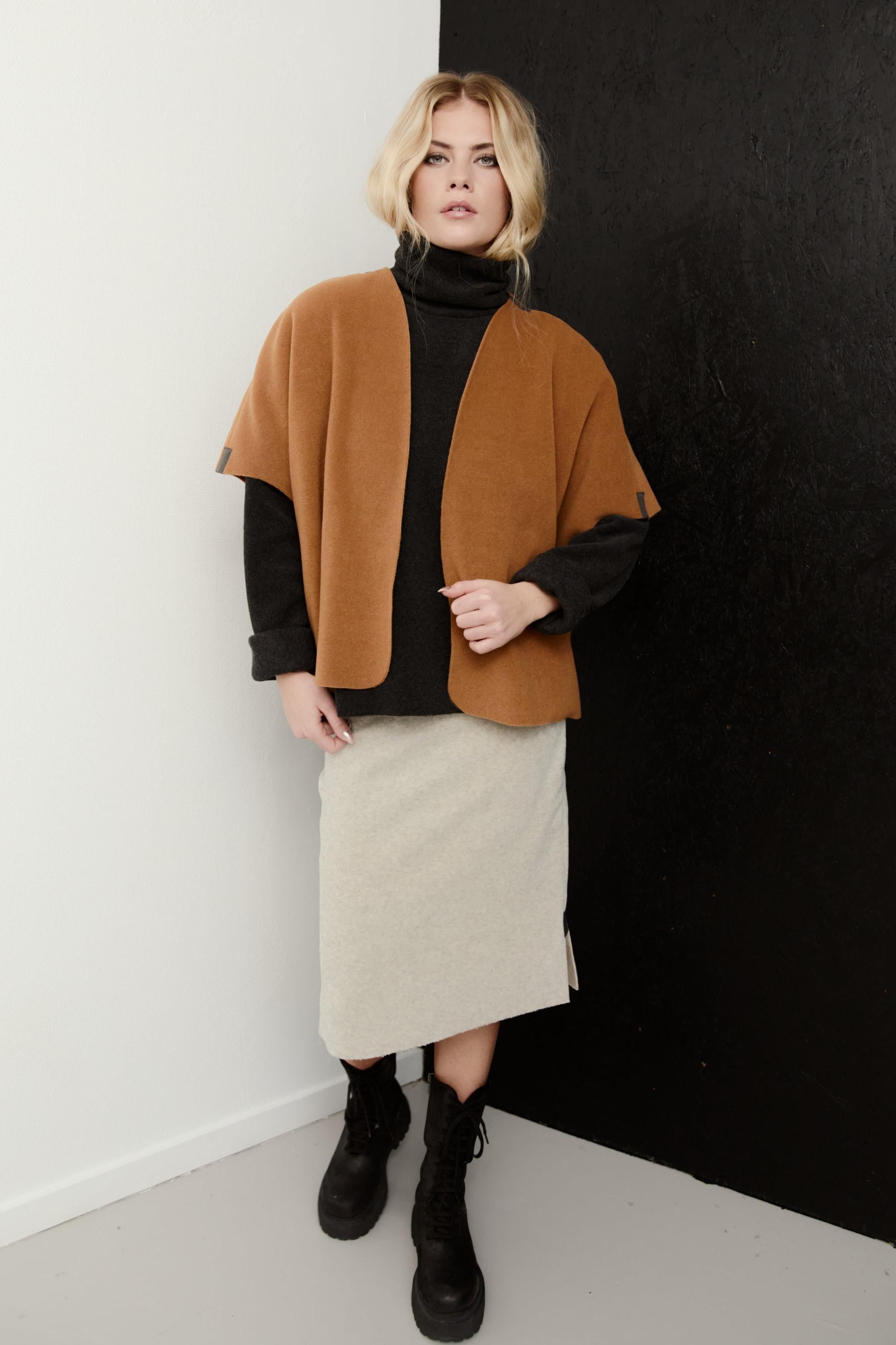 A woman models a cape jacket in camel brown, worn over a black turtleneck and a light beige skirt, standing against a split white and black wall.