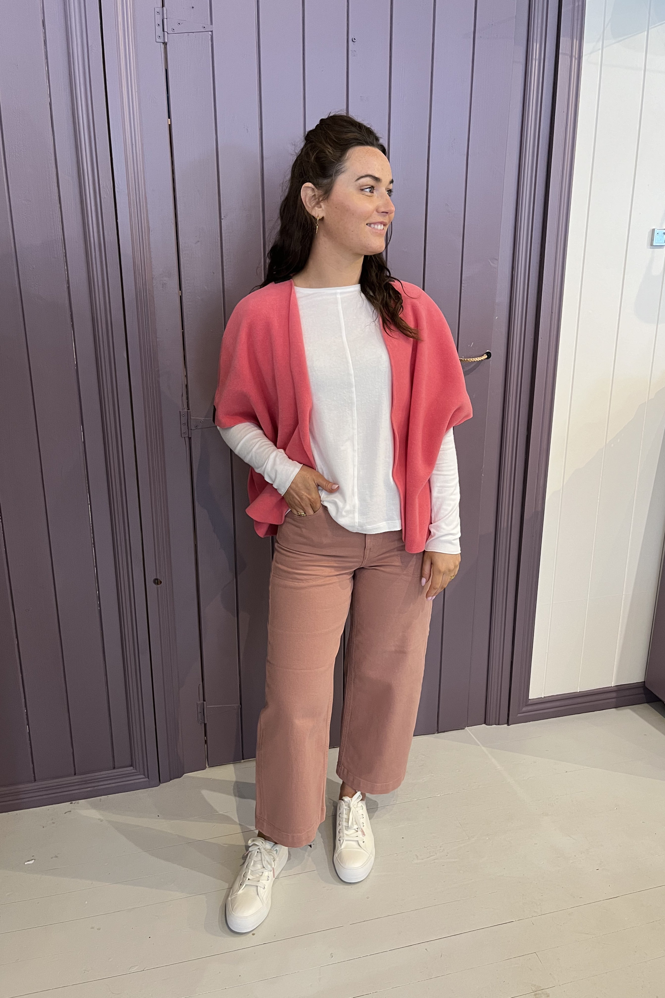 A woman stands indoors wearing a coral pink open-front cardigan over a white long-sleeve top, paired with dusty rose wide-leg pants and white sneakers, posed in front of purple paneled doors.