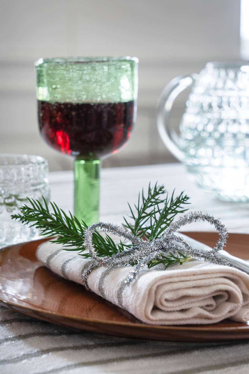 A rustic place setting featuring a linen napkin with a festive pine and silver tinsel ornament on a wooden plate.