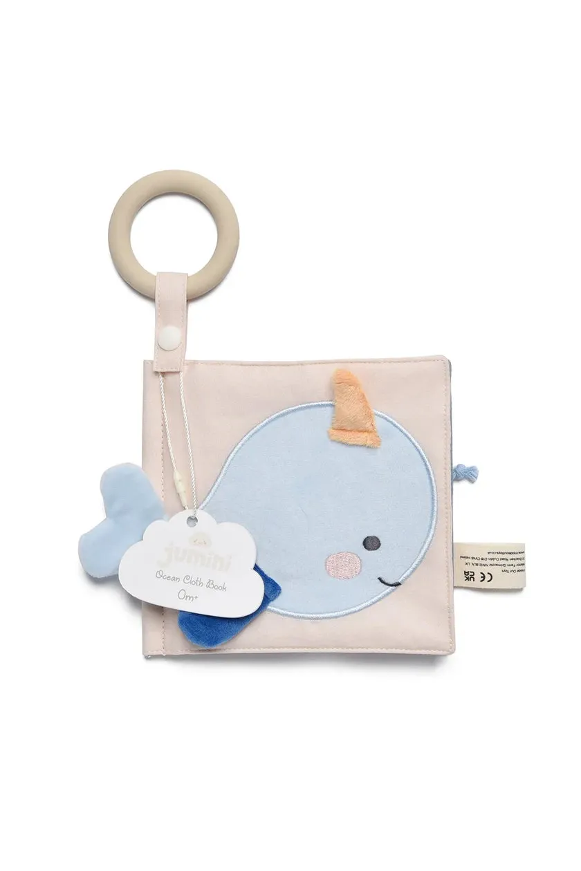 A baby cloth book toy featuring a light blue whale applique on a pale pink fabric cover, with a wooden ring handle, a blue plush whale tail, and a hanging cloud tag that reads 'jumini'.