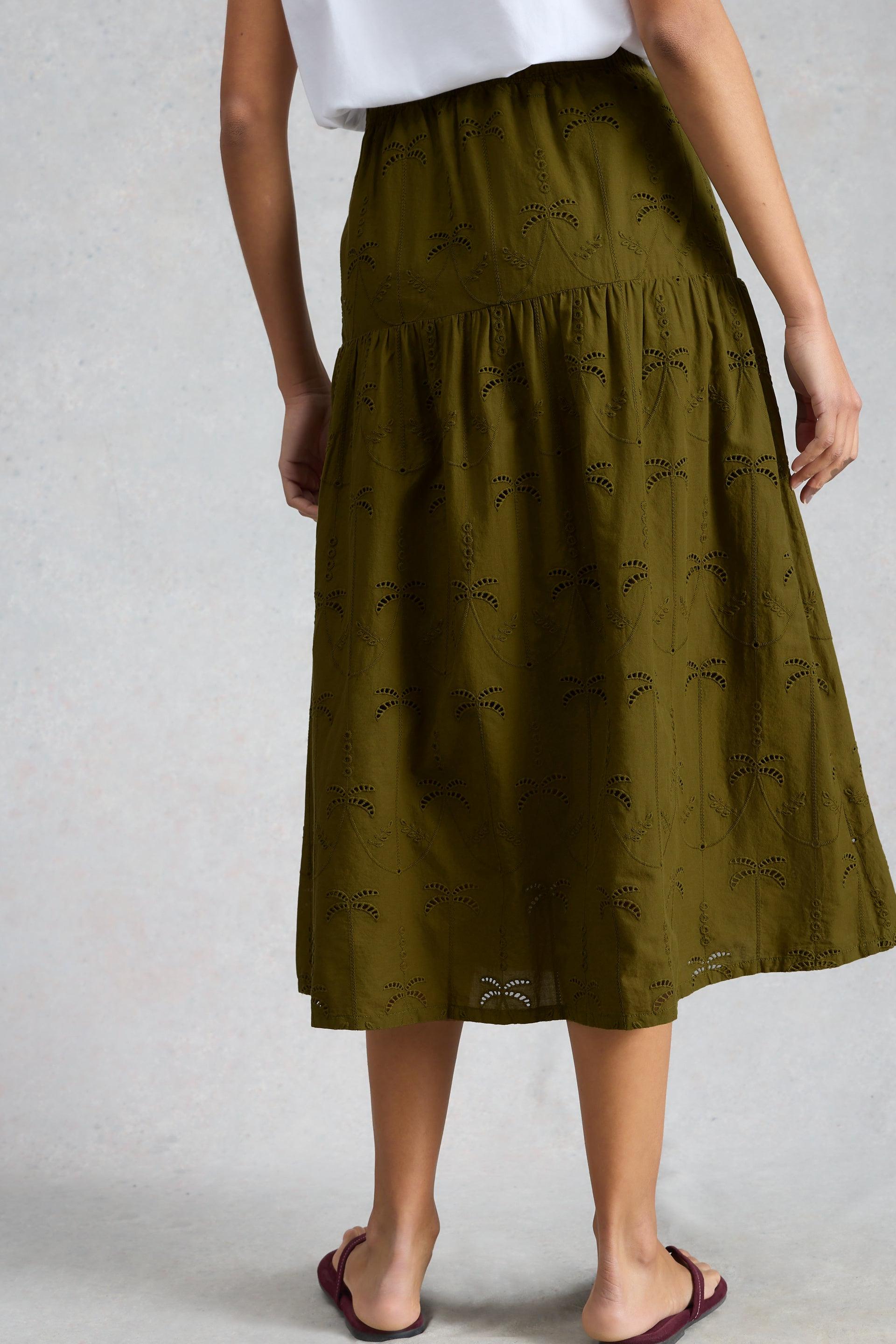 Rear view of a person wearing an olive green organic cotton broderie skirt and simple sandals against a grey background.