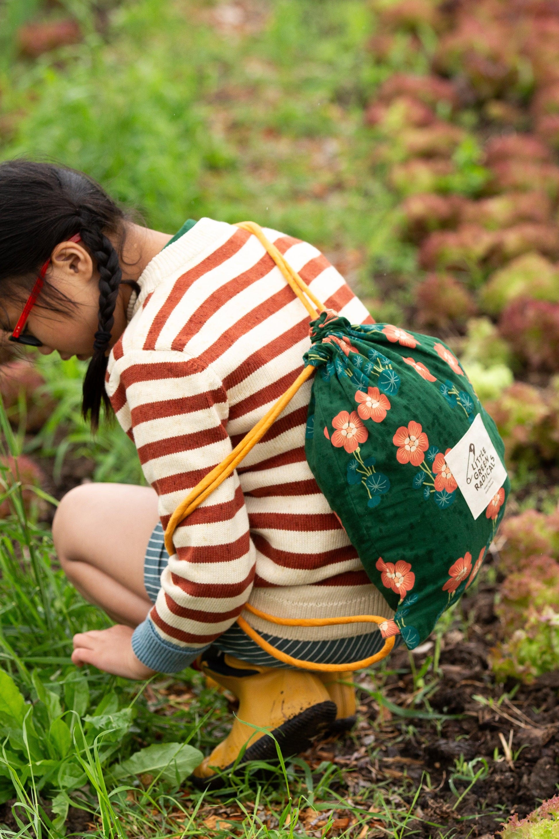 Child in striped organic cotton sweater and floral backpack crouching in a garden