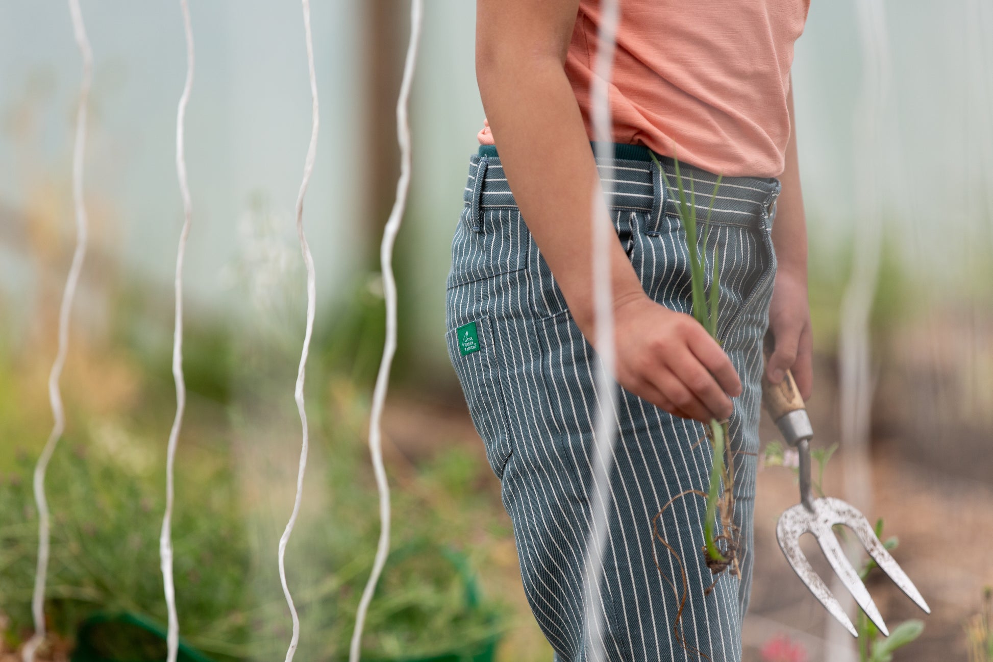 Person in a garden holding a tool, wearing striped pants with a visible Little Green Radicals logo.
