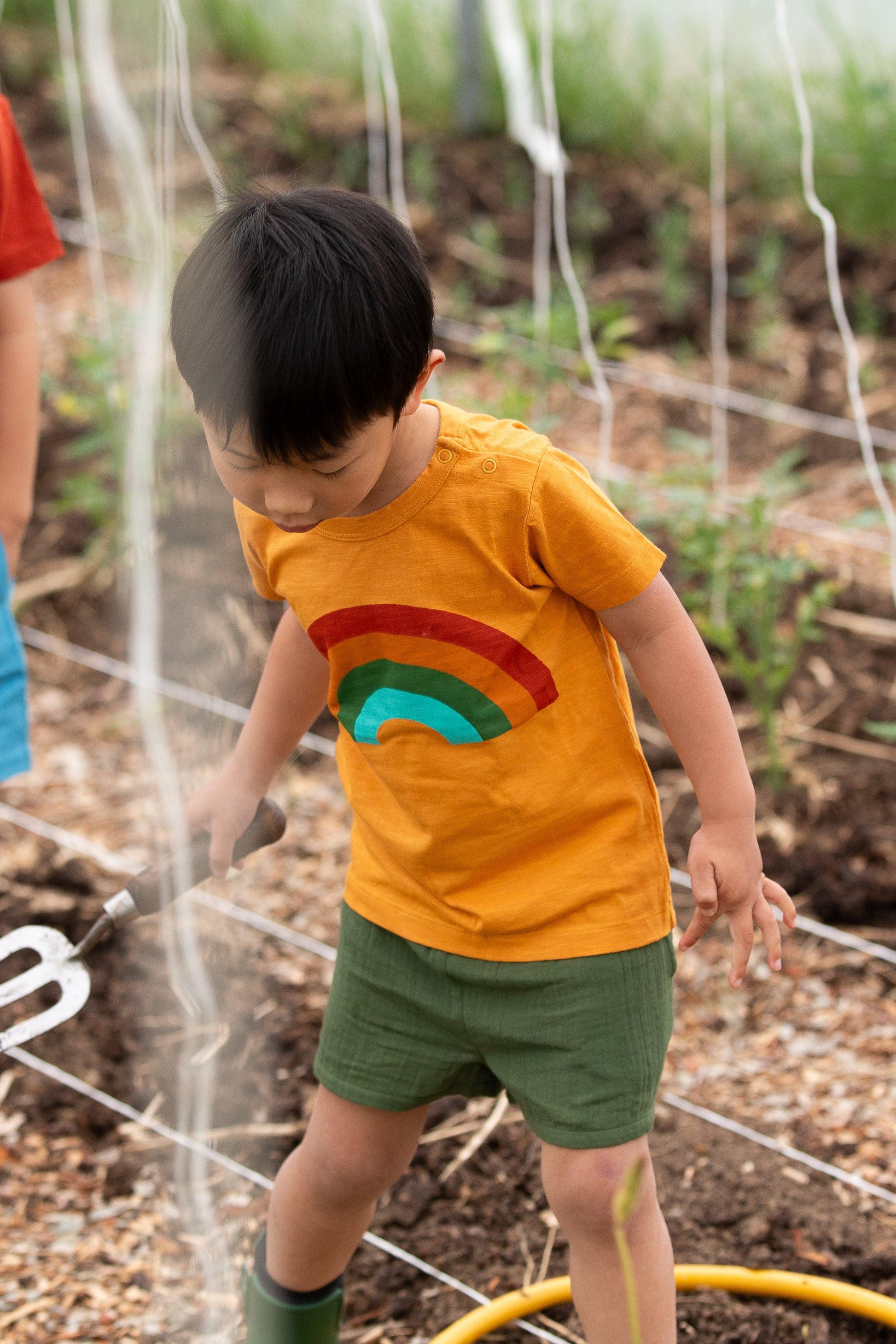 Child wearing a yellow shirt with a rainbow design, standing in a garden.