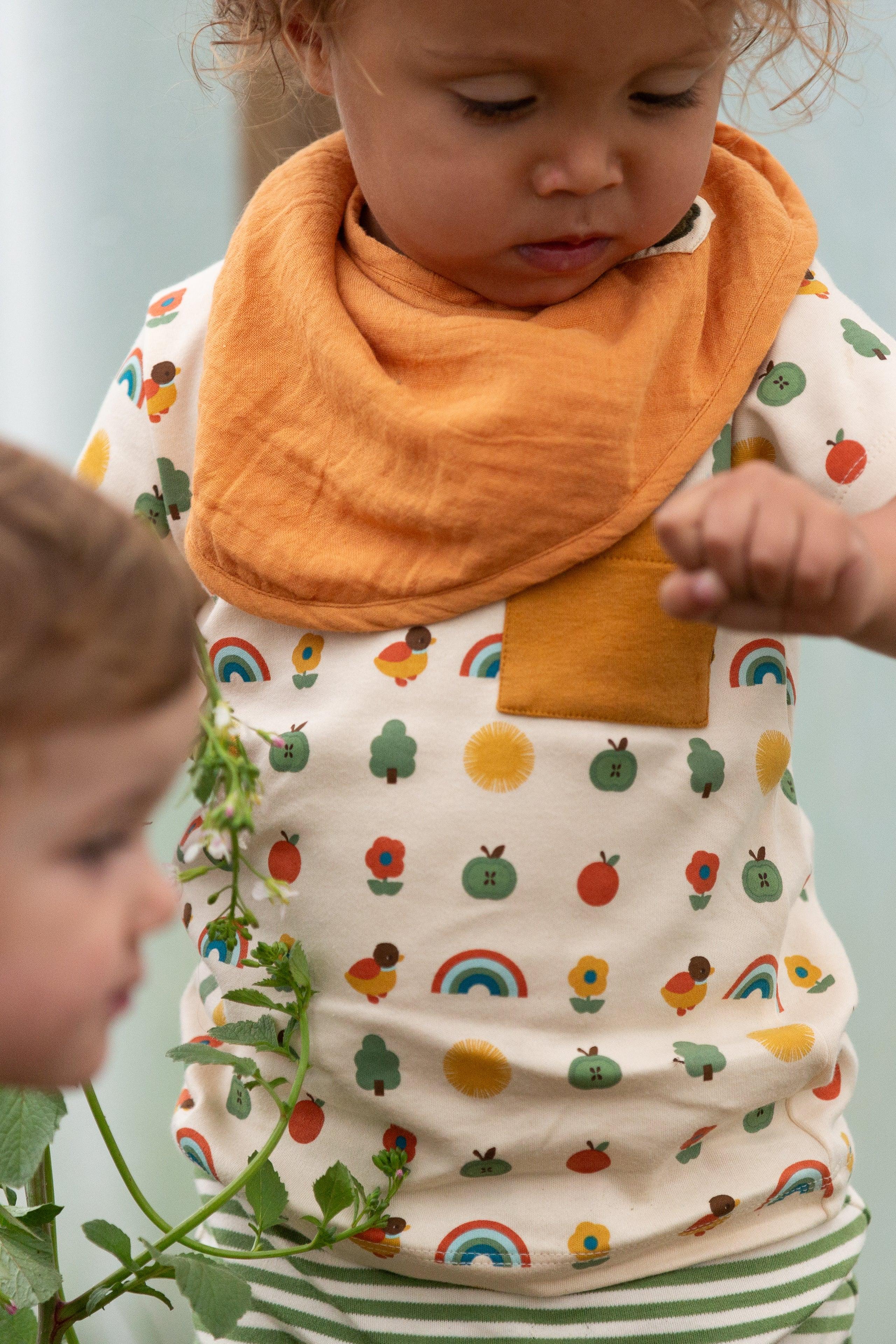Child wearing a colourful bib and organic cotton top with fruit and rainbow pattern, standing outdoors.