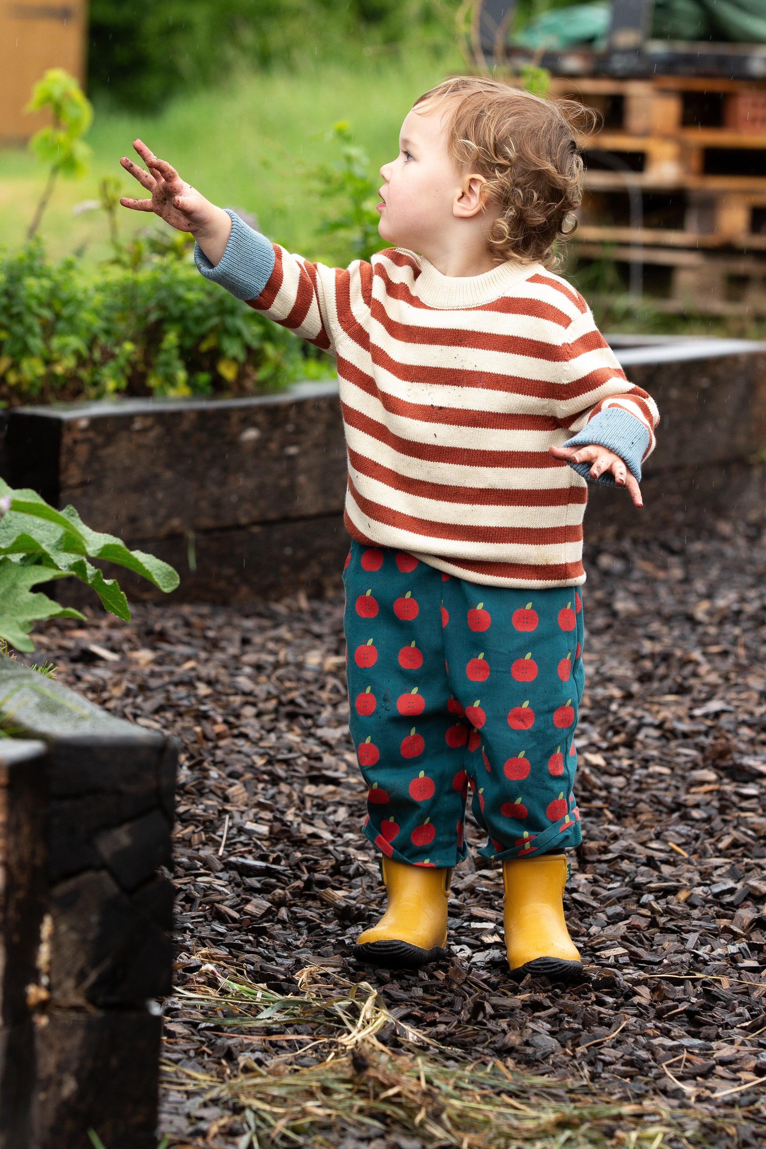 Child in a striped organic cotton sweater and apple print pants standing in a garden