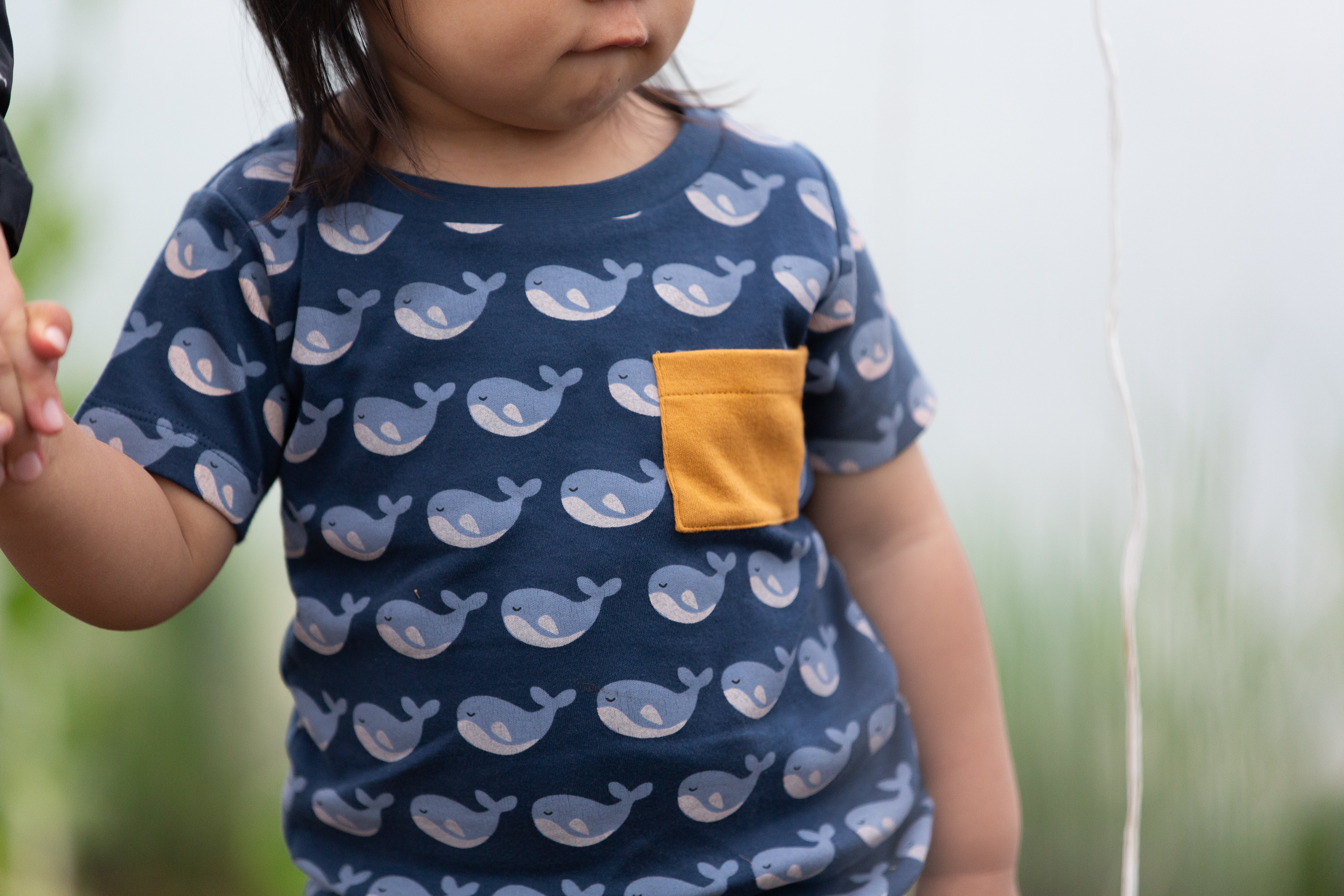 Child wearing a navy blue organic cotton shirt with whale pattern and yellow pocket, standing outdoors.