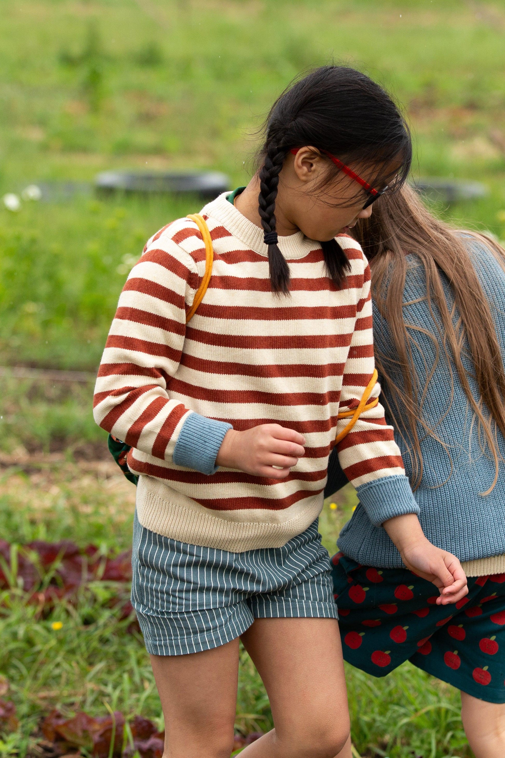 Two children standing in a grassy field, one wearing a striped sweater and shorts, the other in a blue sweater and skirt.