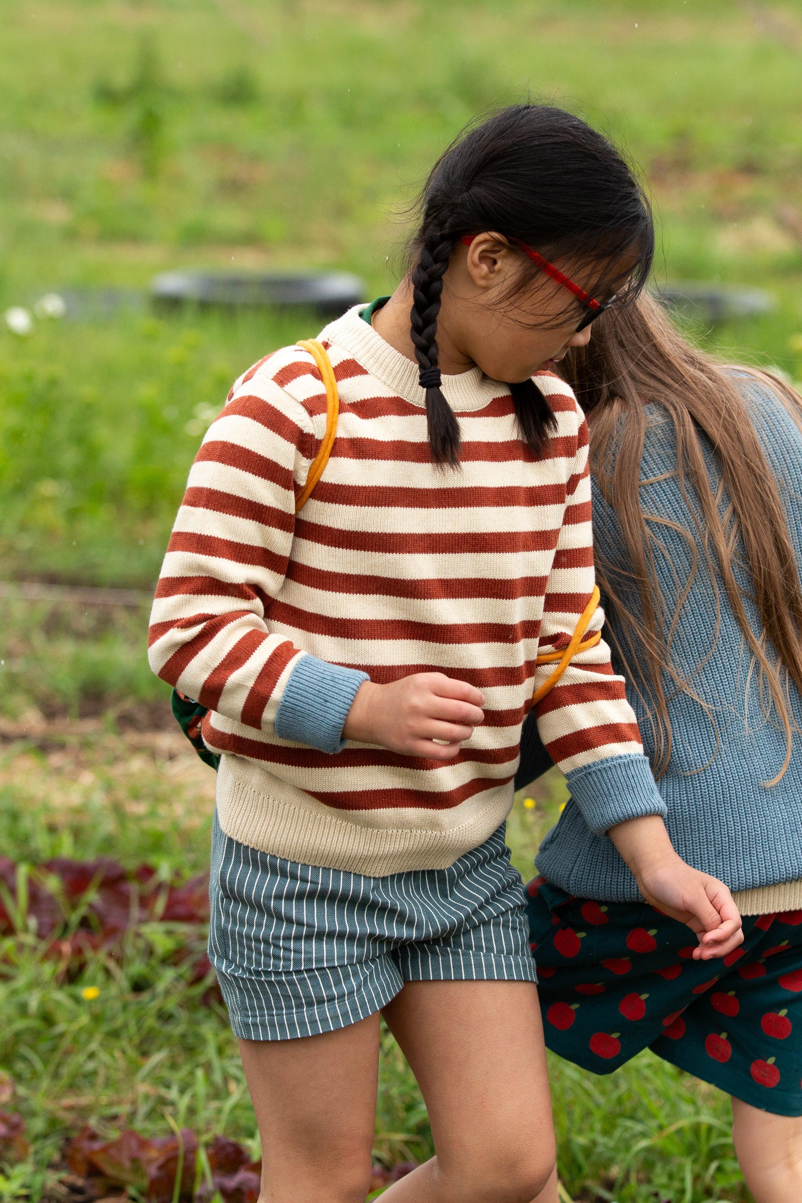 Two children standing in a grassy field, one wearing a striped sweater and shorts, the other in a blue sweater and skirt.