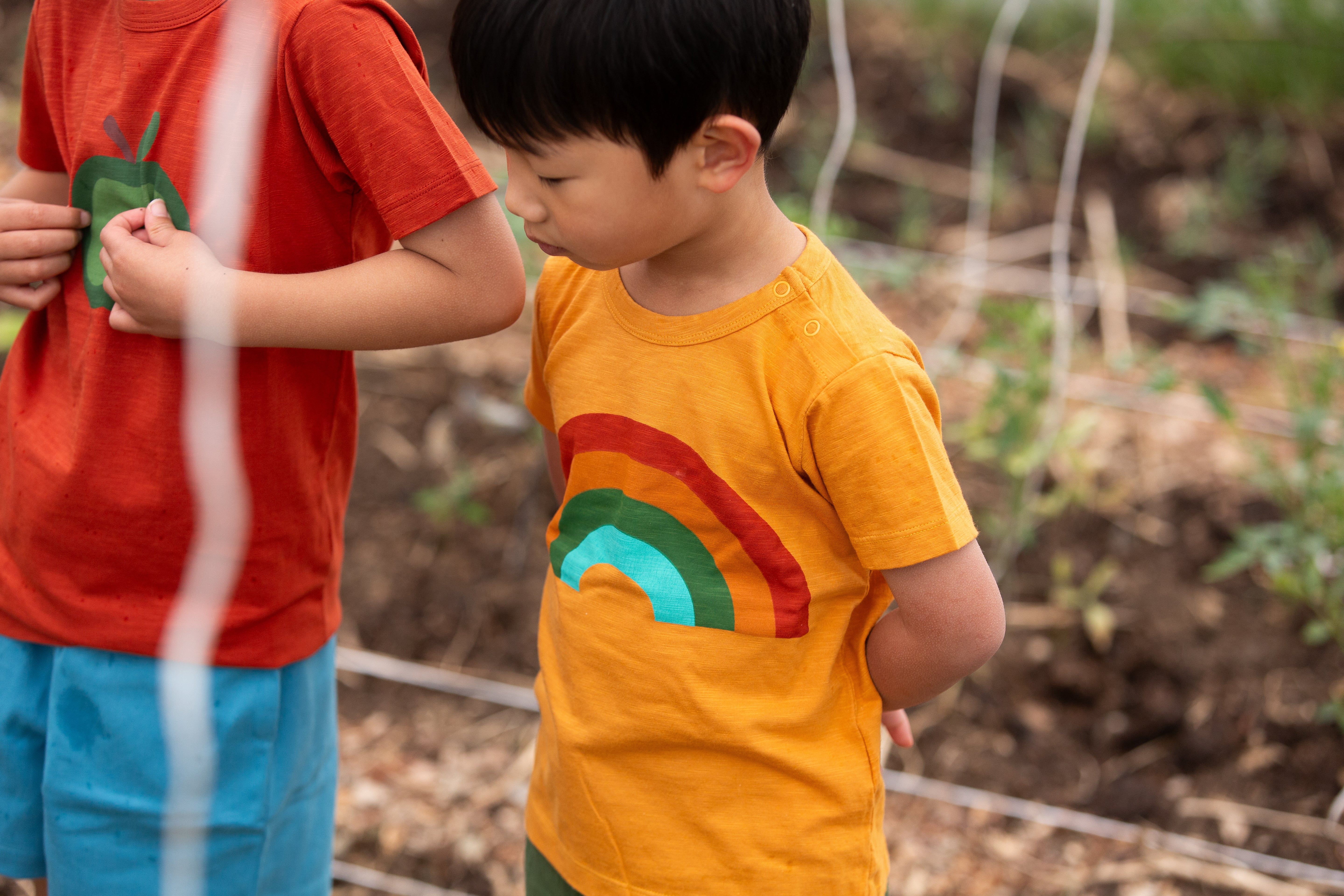 Two children, one in a red shirt and blue shorts, the other in a yellow shirt with a rainbow design, standing in a garden.
