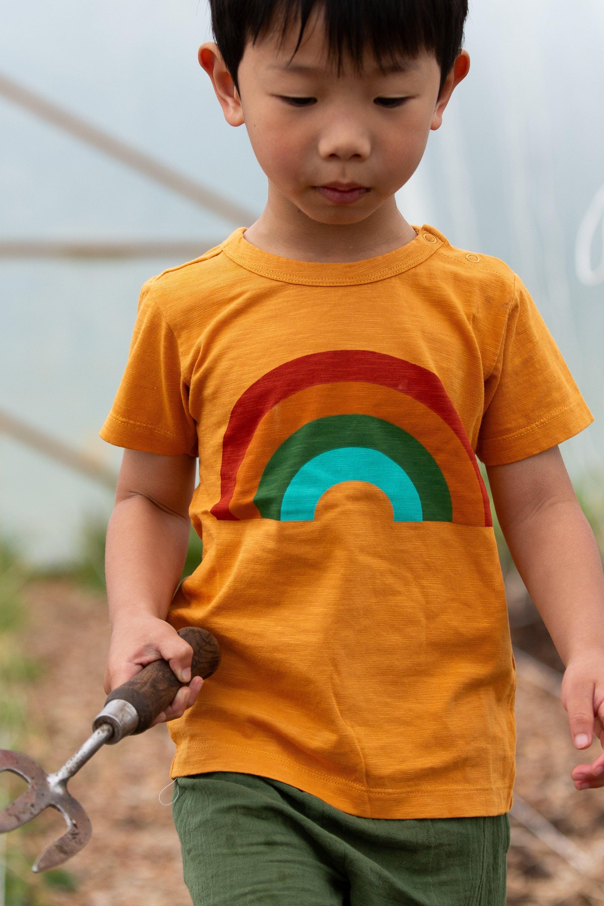 Child wearing an organic cotton rainbow shirt holding a gardening tool in a garden setting