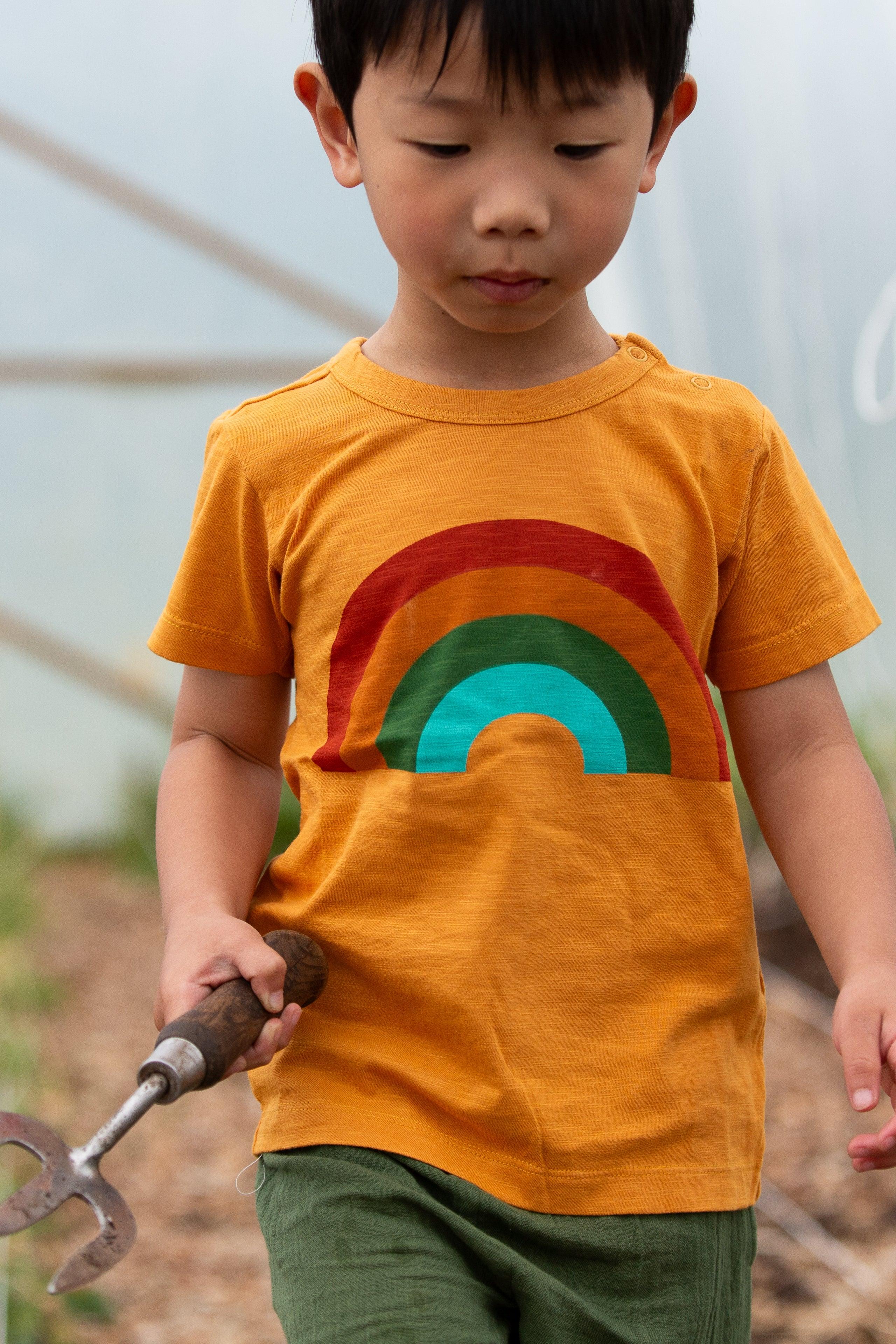 Child wearing an organic cotton rainbow shirt holding a gardening tool in a garden setting