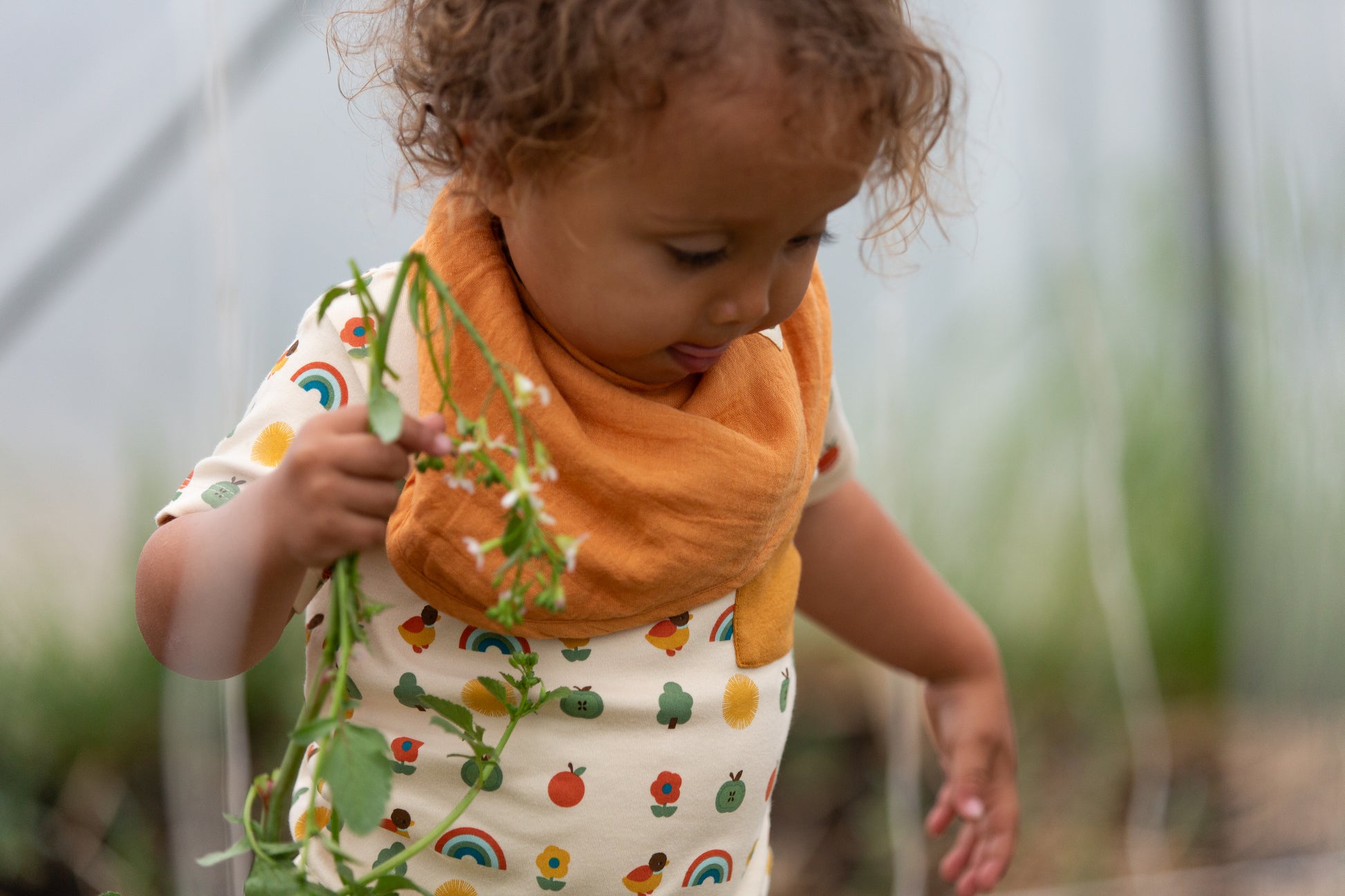 Child wearing an orange bib and patterned organic cotton shirt holding a plant in a garden setting