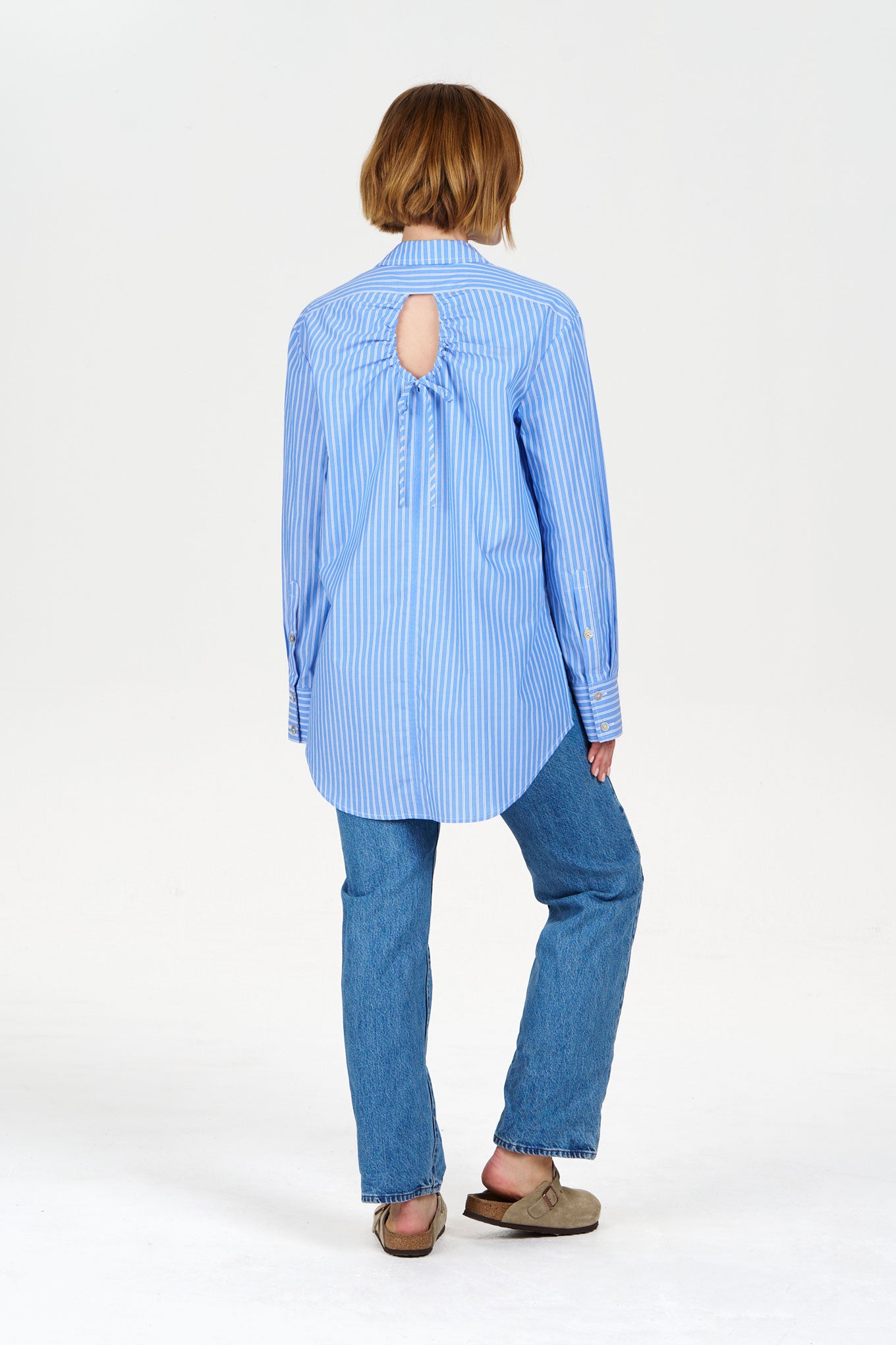 Back view of model wearing a blue striped shirt with hole detail and blue slim jeans on a white background