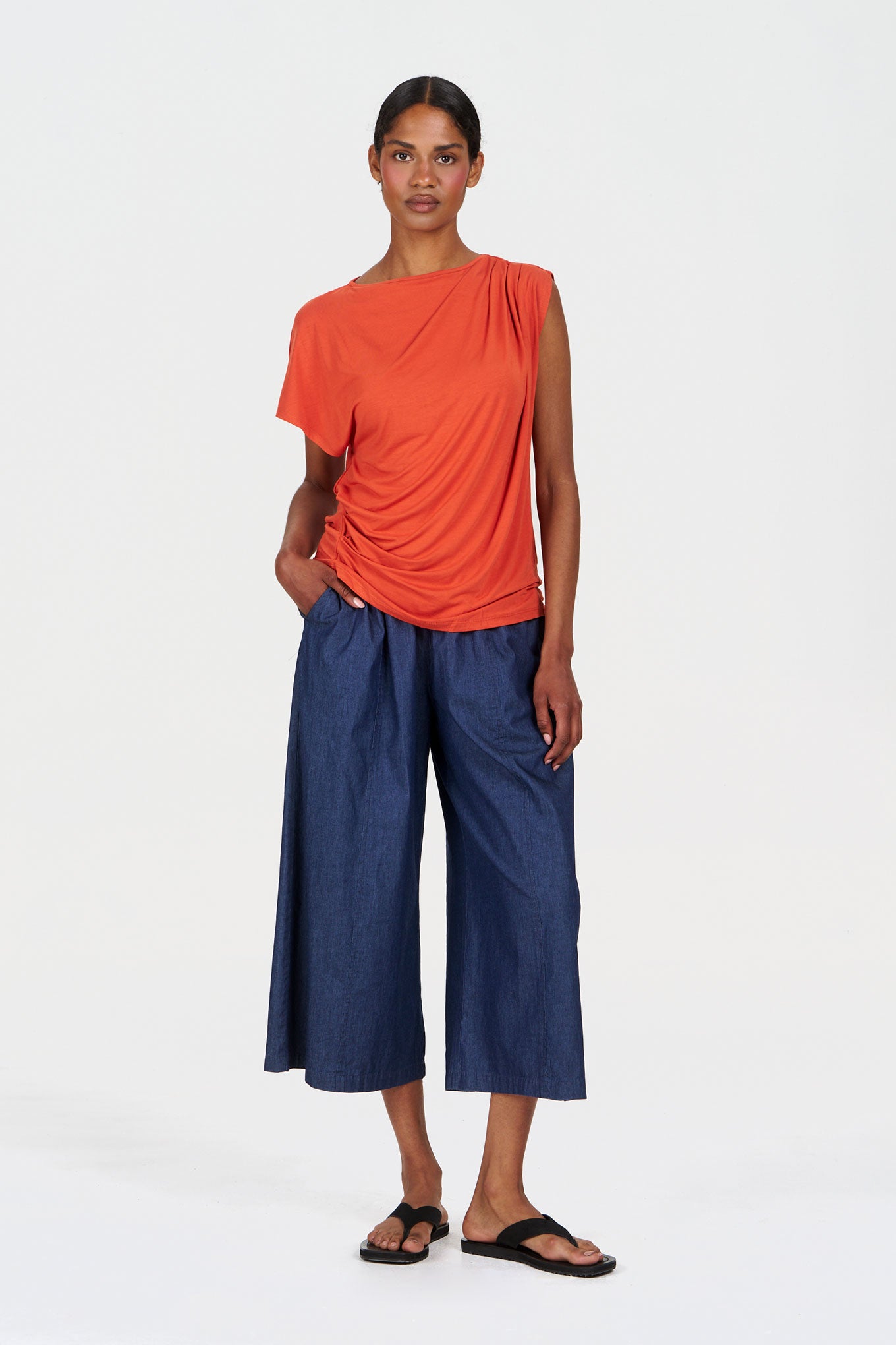 Woman wearing an orange draped top and blue pants on a white background