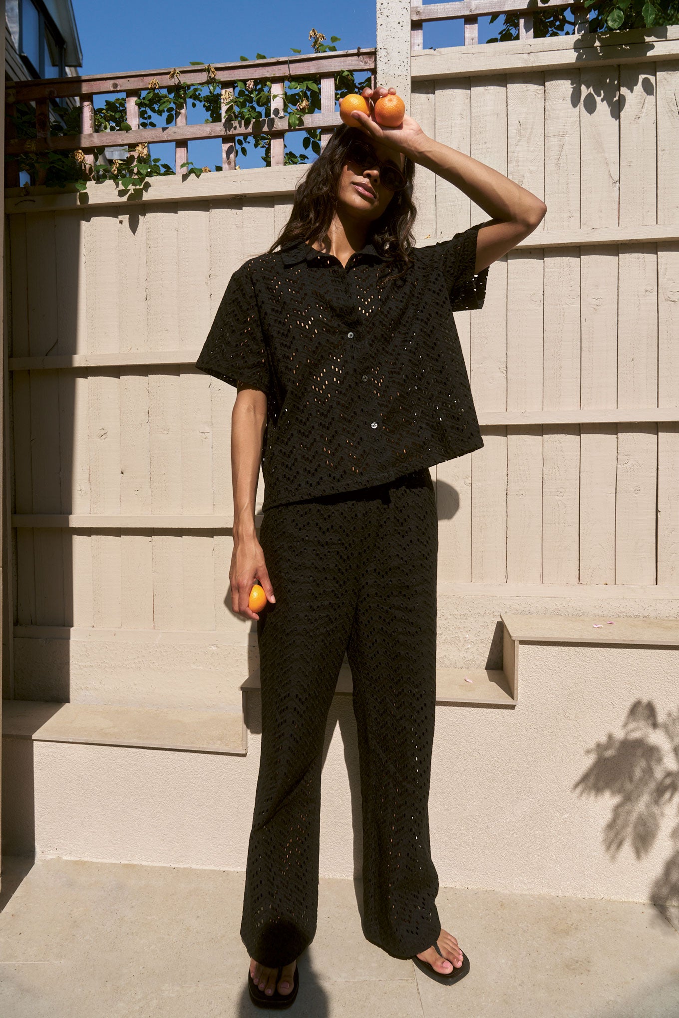 Person wearing a black broderie outfit with a textured pattern, standing outdoors against a wooden fence.