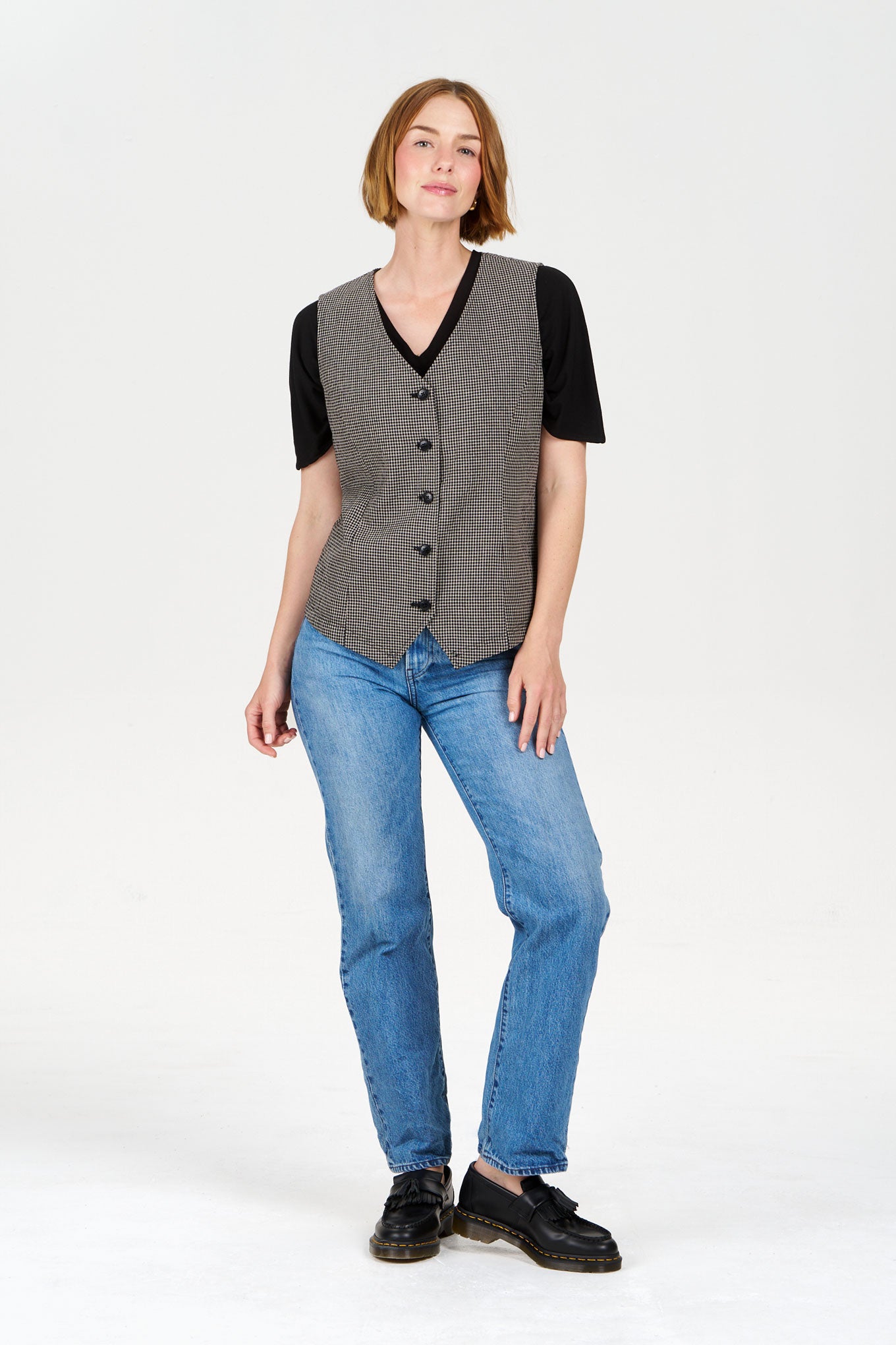 Woman wearing a checkered vest over a black shirt with blue jeans on a white background
