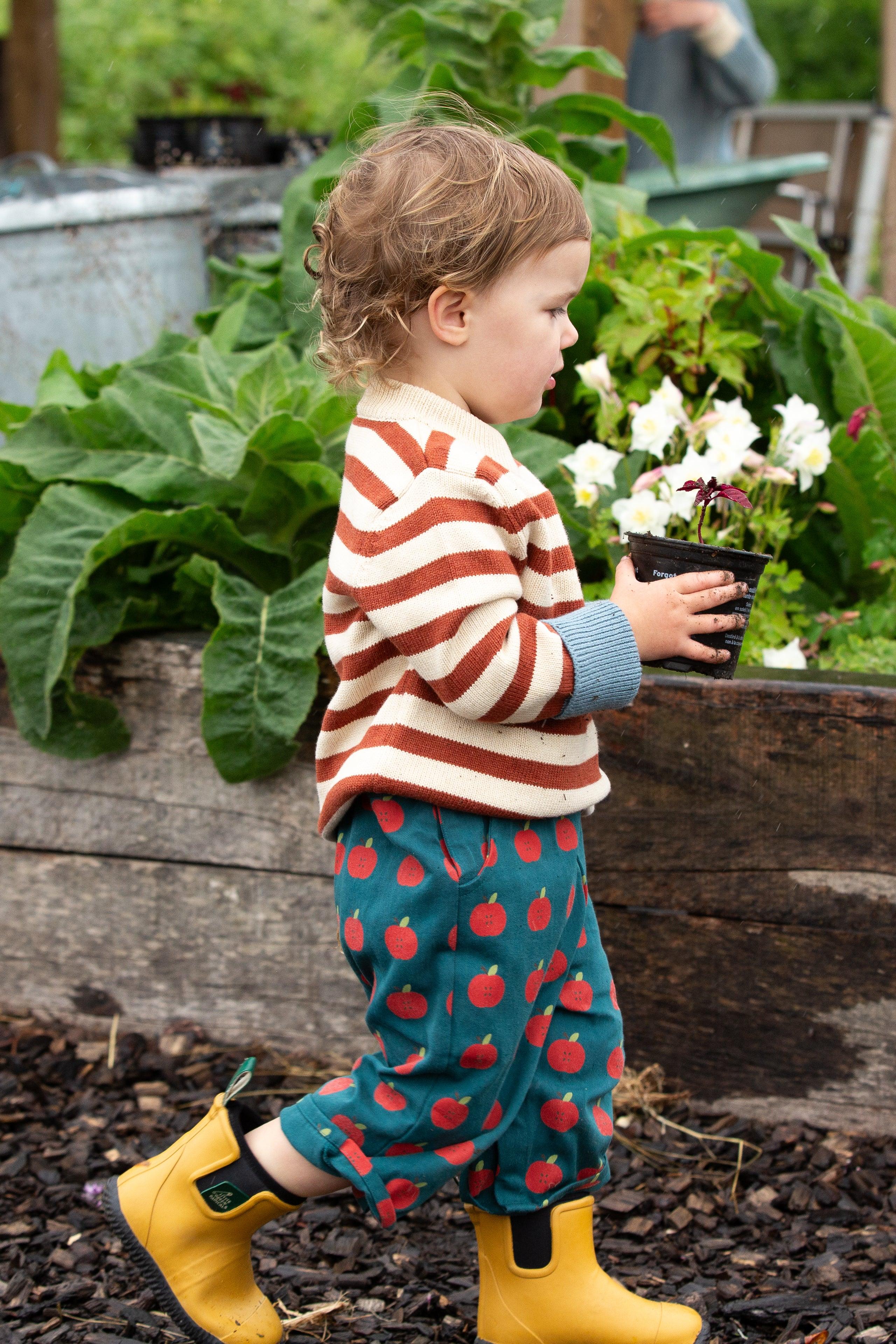 Child in a garden holding a potted plant, wearing a striped knit sweater and yellow rain boots.