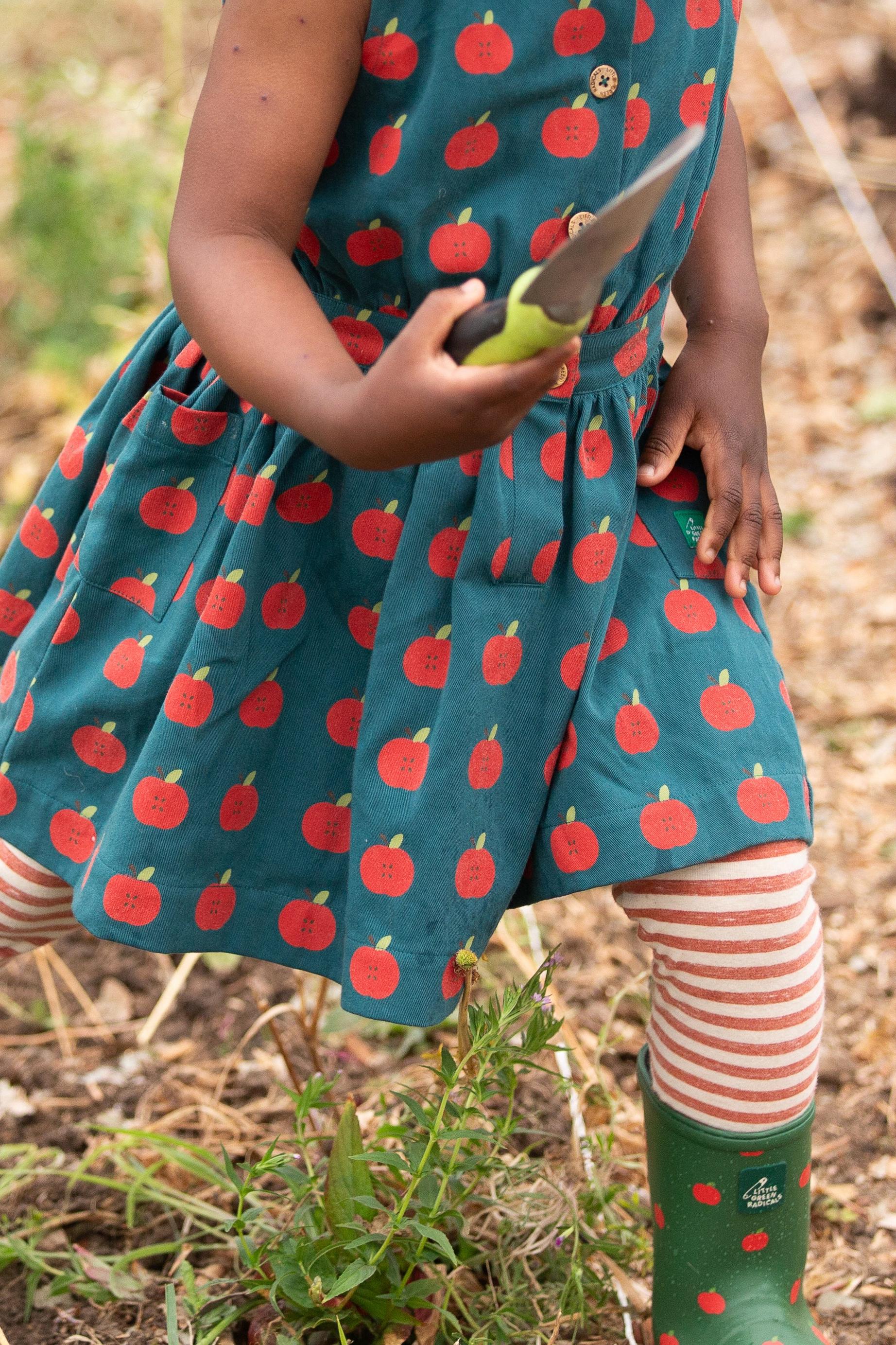 Child wearing a dress with apple pattern, striped leggings and holding gardening tools outdoors