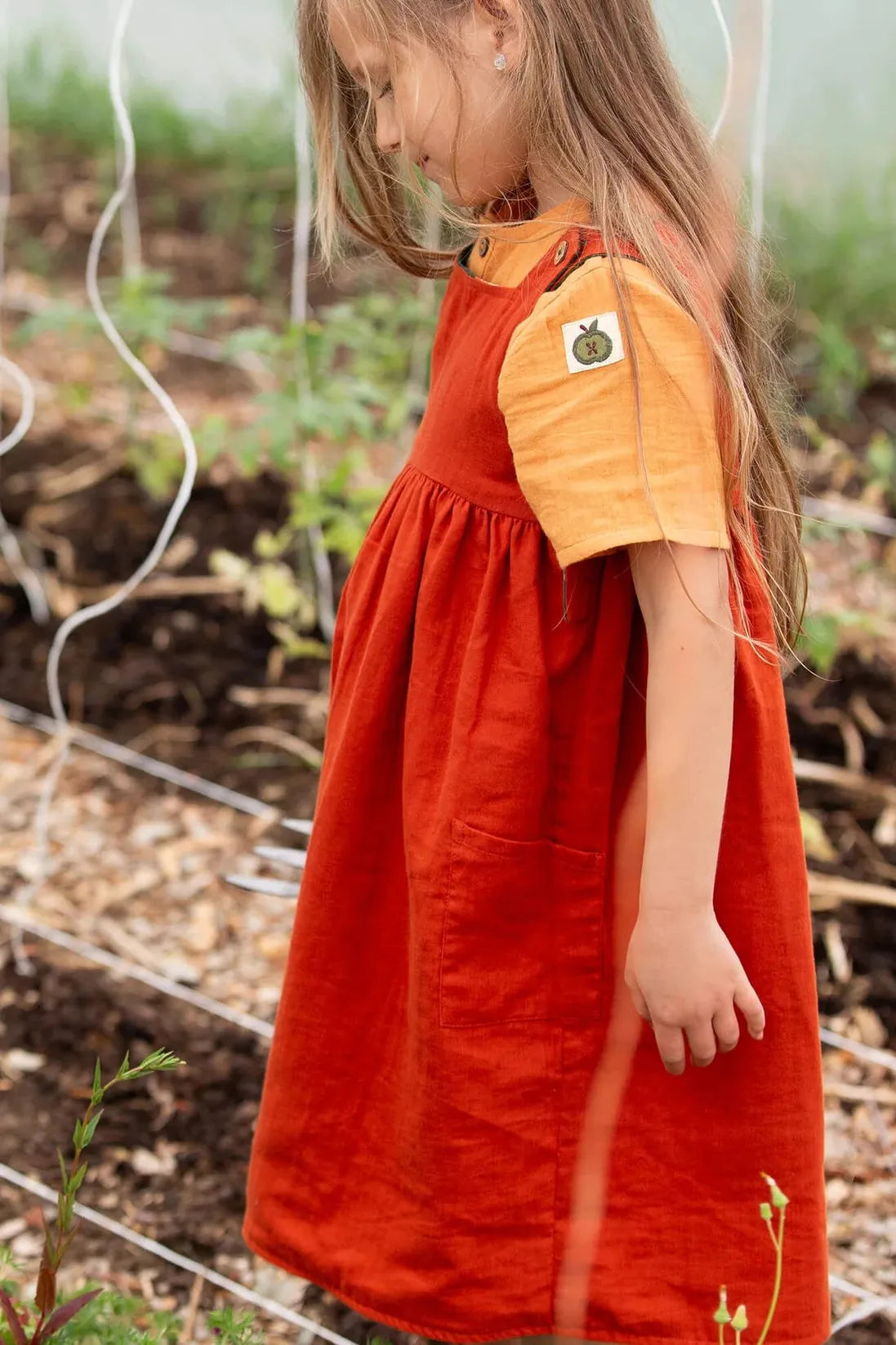 Child in a red organic cotton dress with an orange shirt standing in a garden