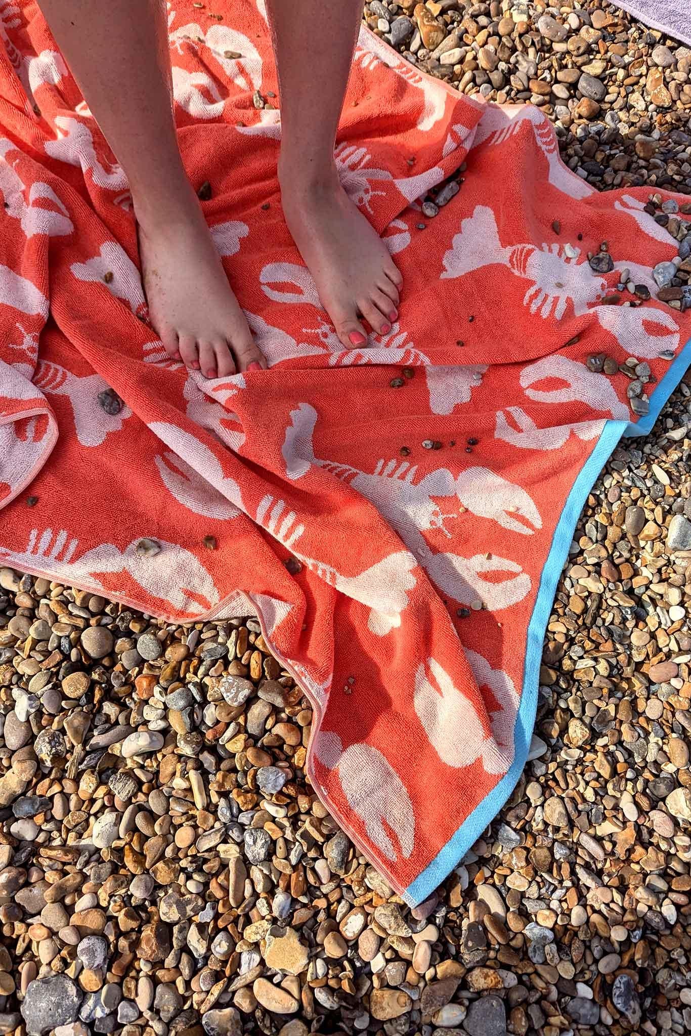 Person standing on a red towel with lobster pattern on pebbly ground