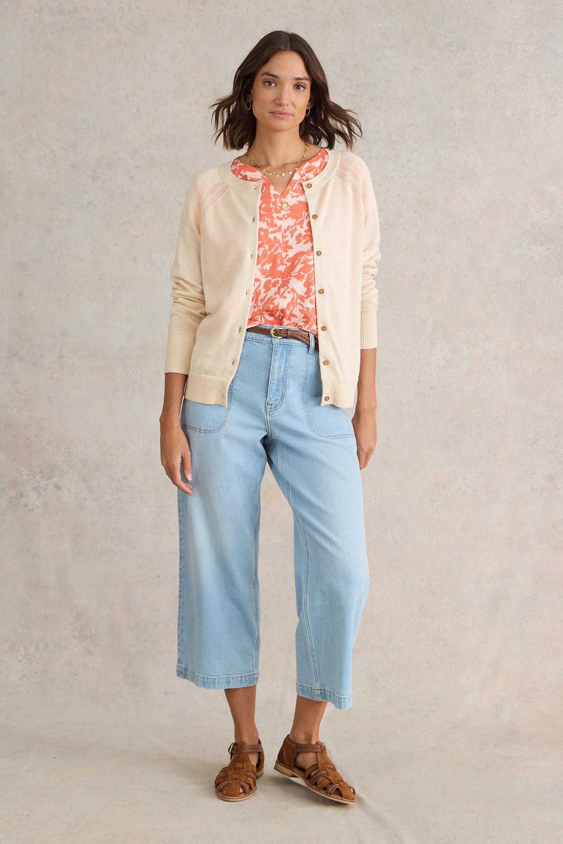 Woman wearing a beige organic cotton cardigan, coral top, light blue jeans, and brown sandals on a neutral background