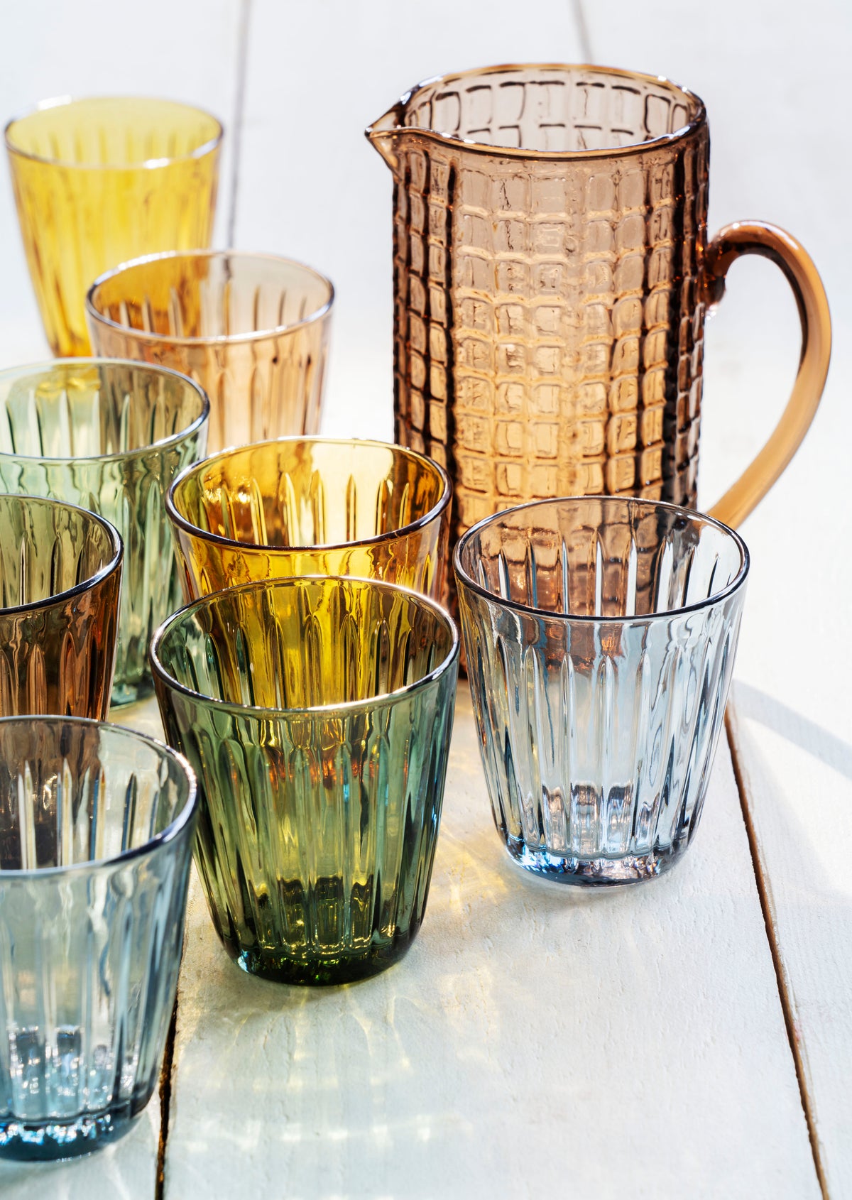 A collection of colourful Madam Stoltz drinking glasses and a matching glass pitcher on a white wooden table.