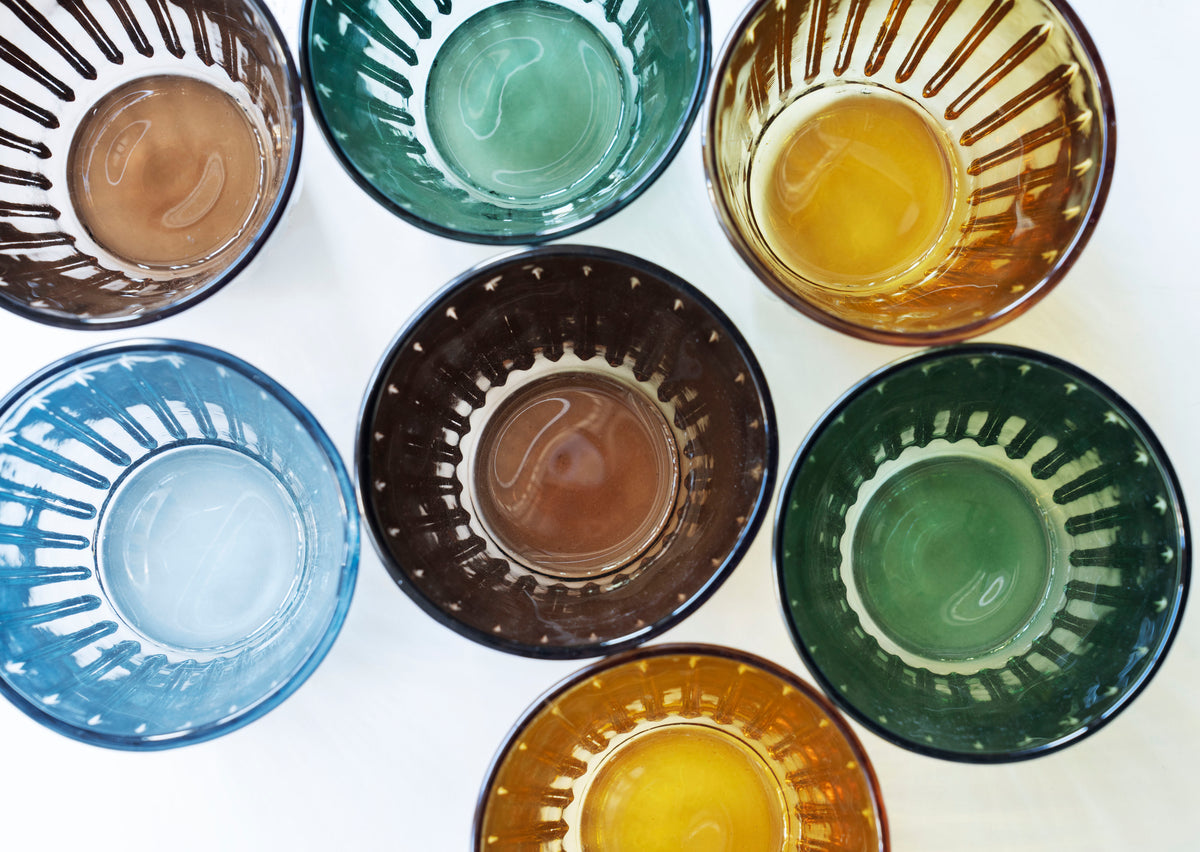 Top-down view of seven colourful, ribbed Madam Stoltz drinking glasses arranged on a white surface.