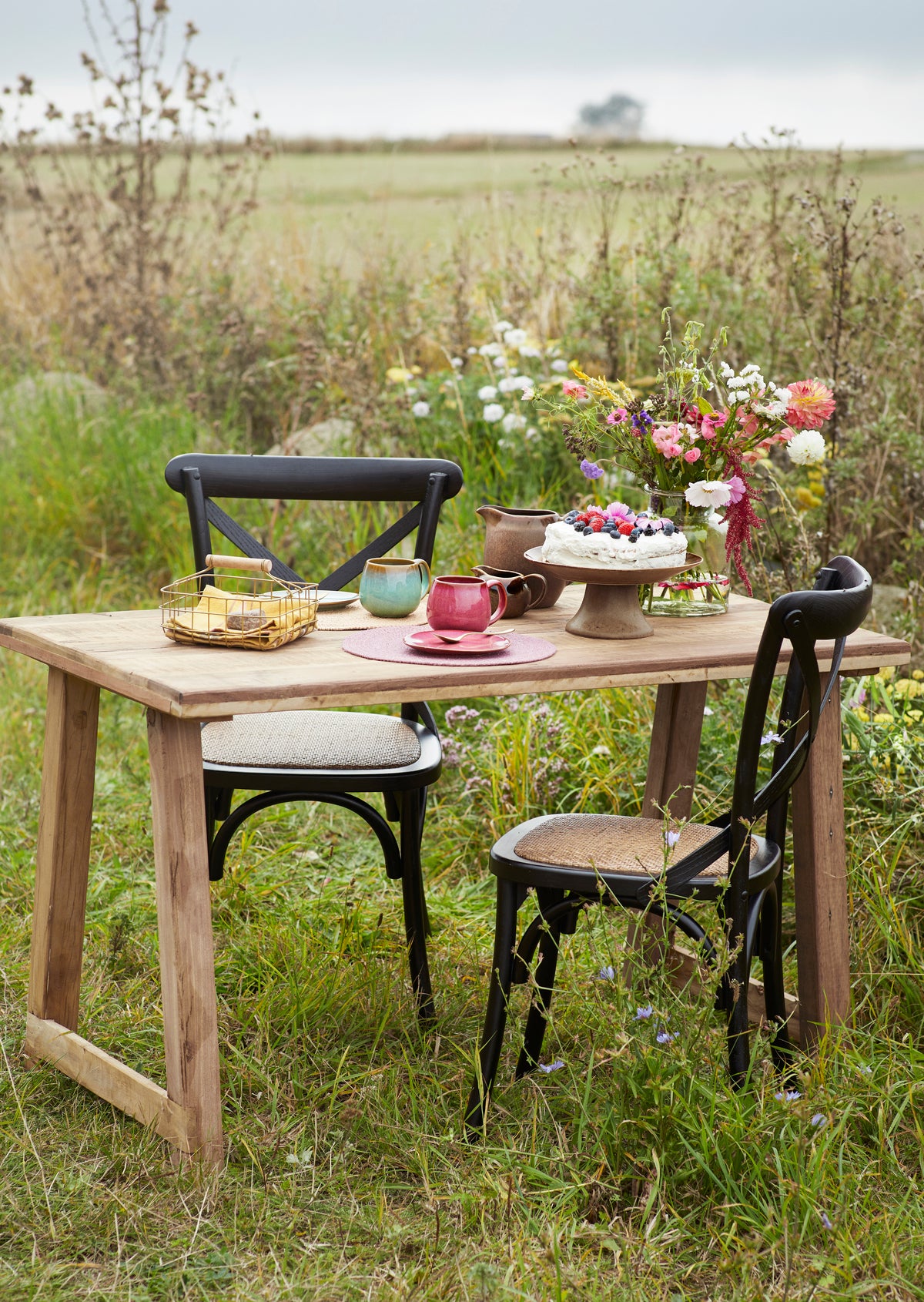 A Madam Stoltz plum ceramic coffee mug on a wooden table with cake and a bouquet in a meadow.