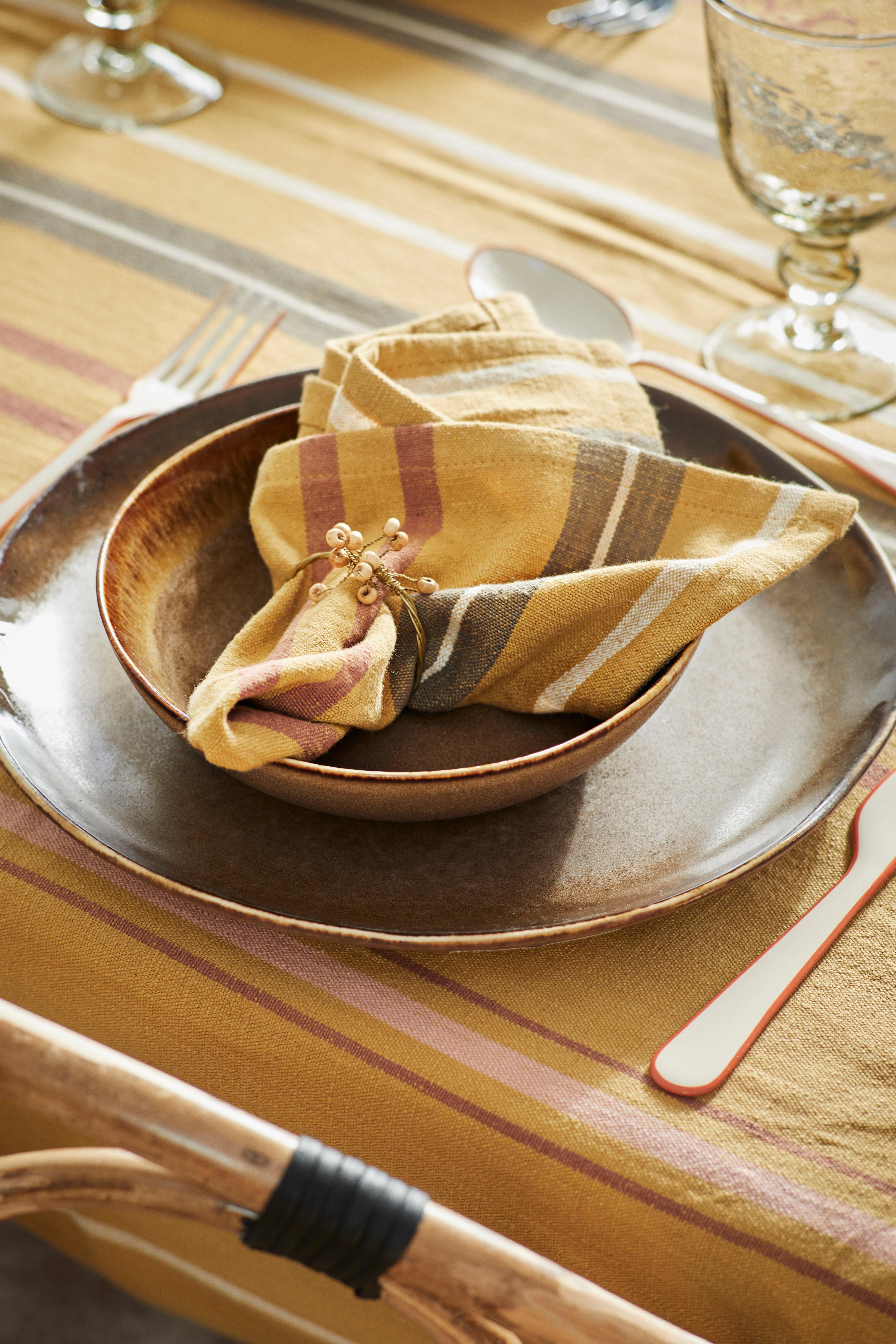 Dinner setting with a gold plate, striped napkin, and cutlery on a striped tablecloth.