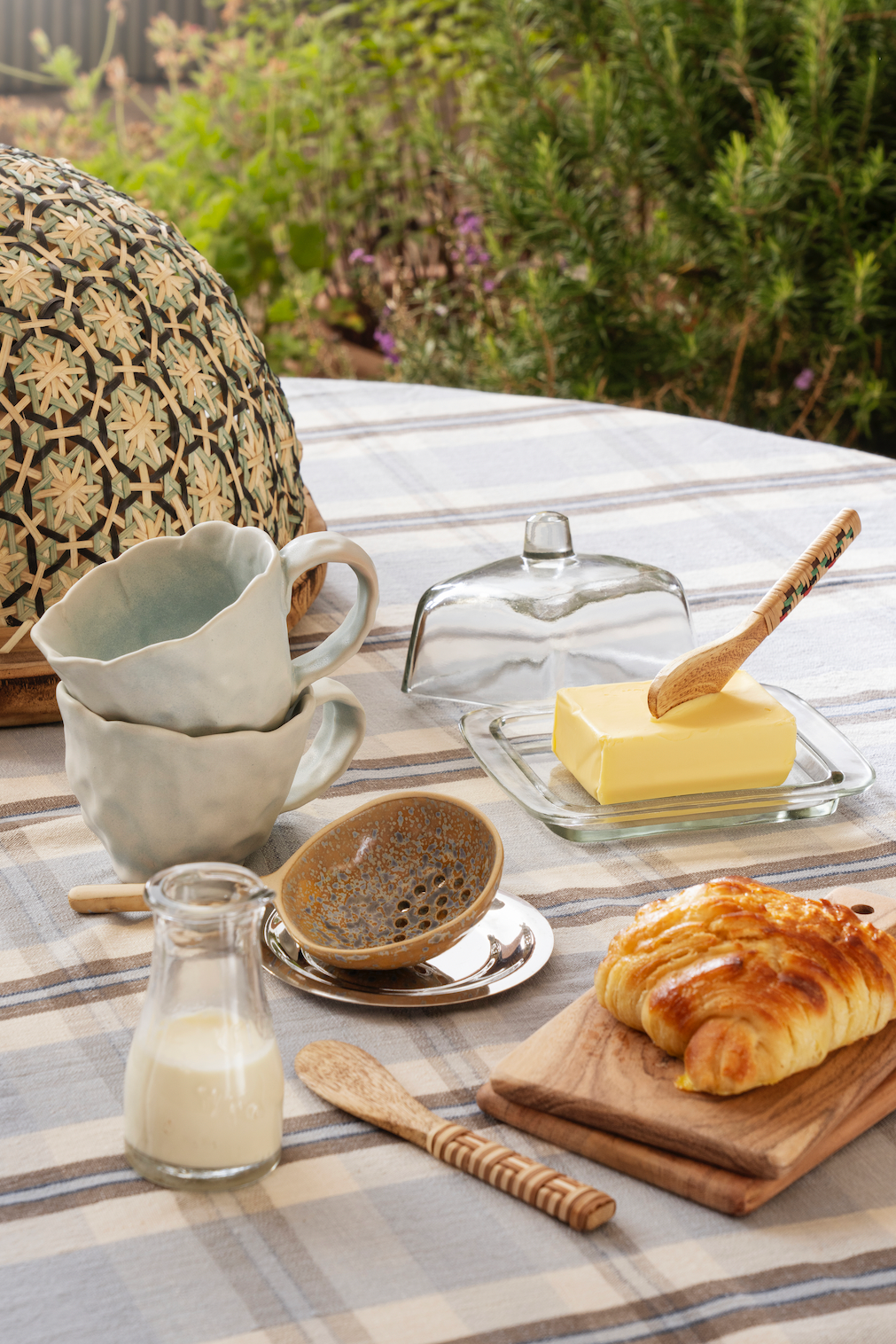 Breakfast setting with croissant, butter, and coffee on a plaid tablecloth outdoors.